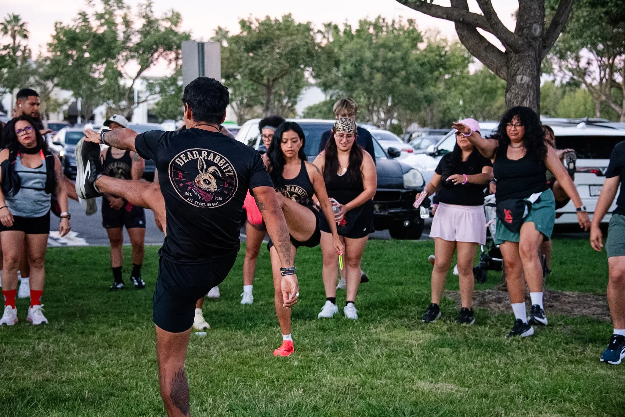 A group of people participating in an outdoor fitness class or dance session in a park, led by an instructor in a black t-shirt.