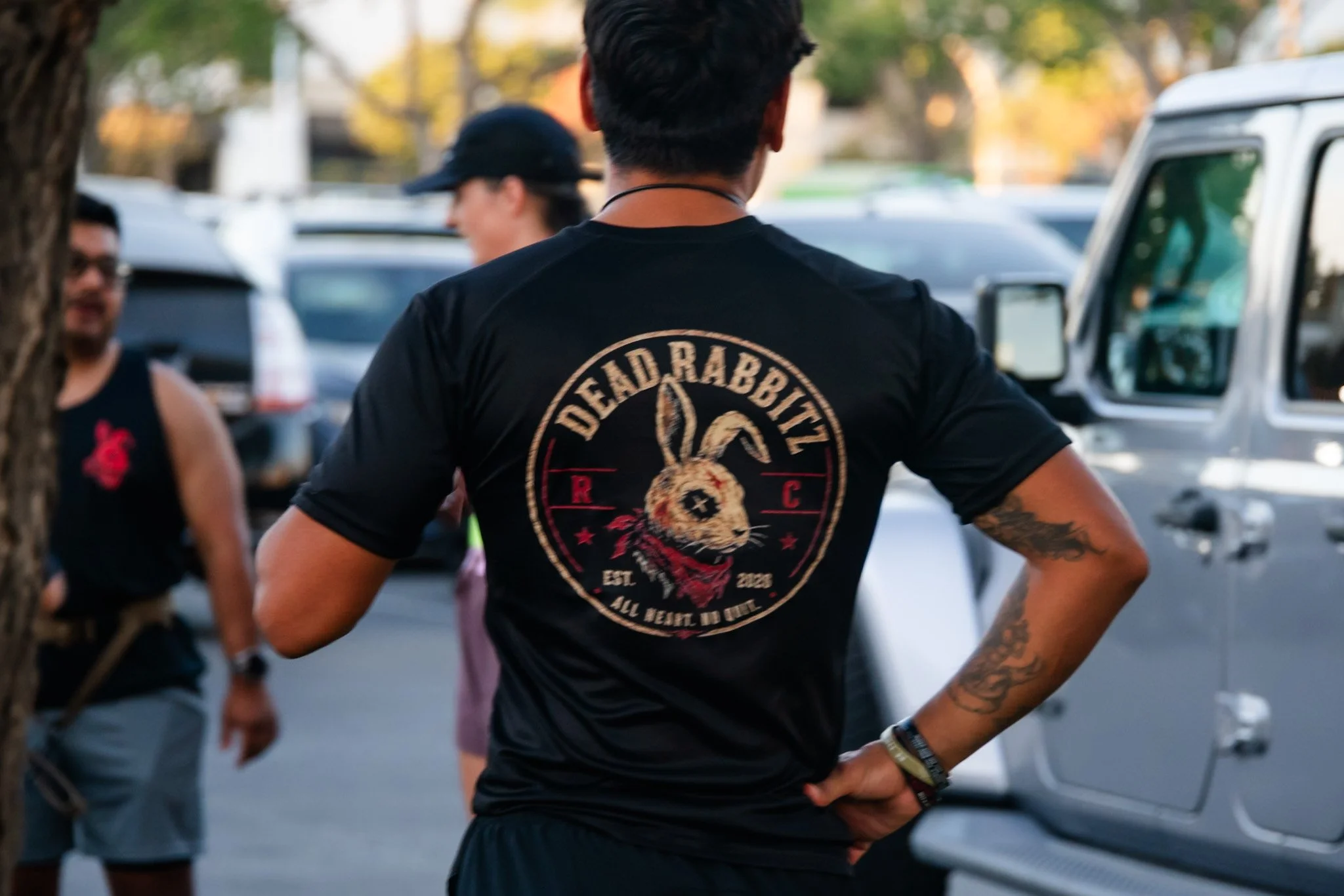 A person with short dark hair, tattoos on their arm, and a black t-shirt with a graphic of a rabbit and the words "Deadrabbit" seen from the back, standing outdoors near parked cars and talking to other people.
