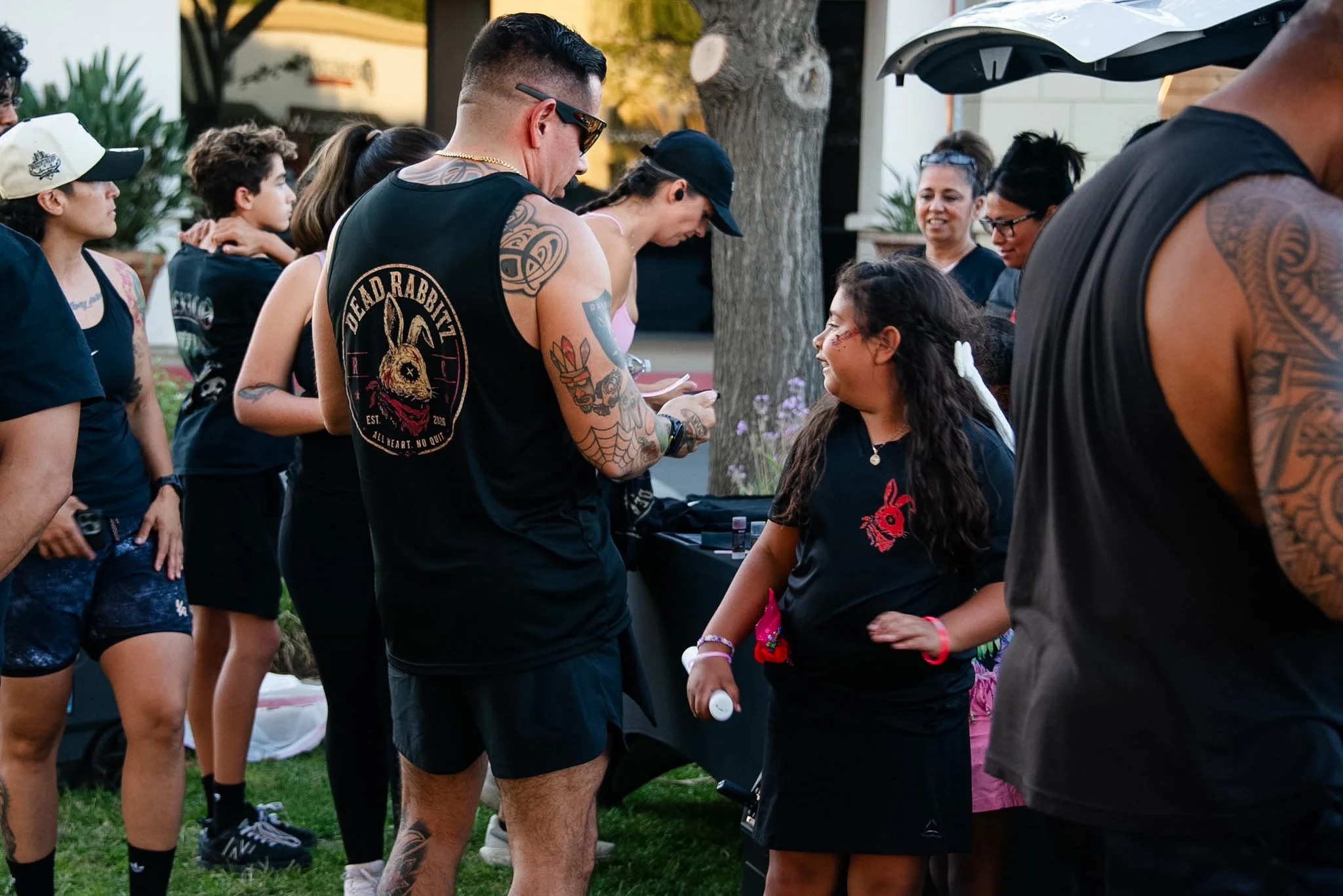 A group of people lined up outdoors at a gathering, engaging in conversation. The group includes adults and children, some wearing tattoos, hats, and casual summer clothing, standing near a table with items on it.