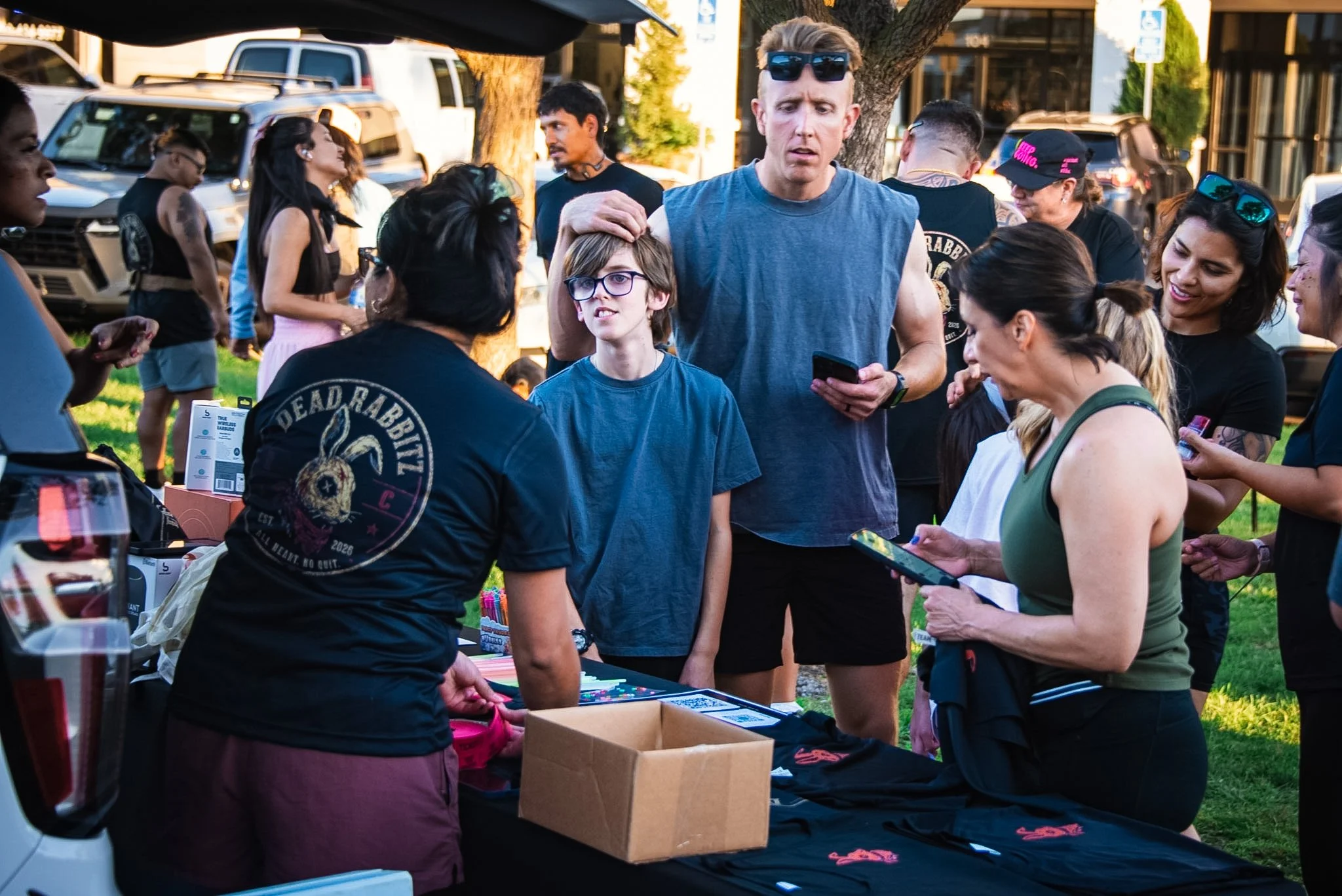 People gathered outdoors around a table at a community event, engaging in conversations, shopping, and socializing on a sunny day.