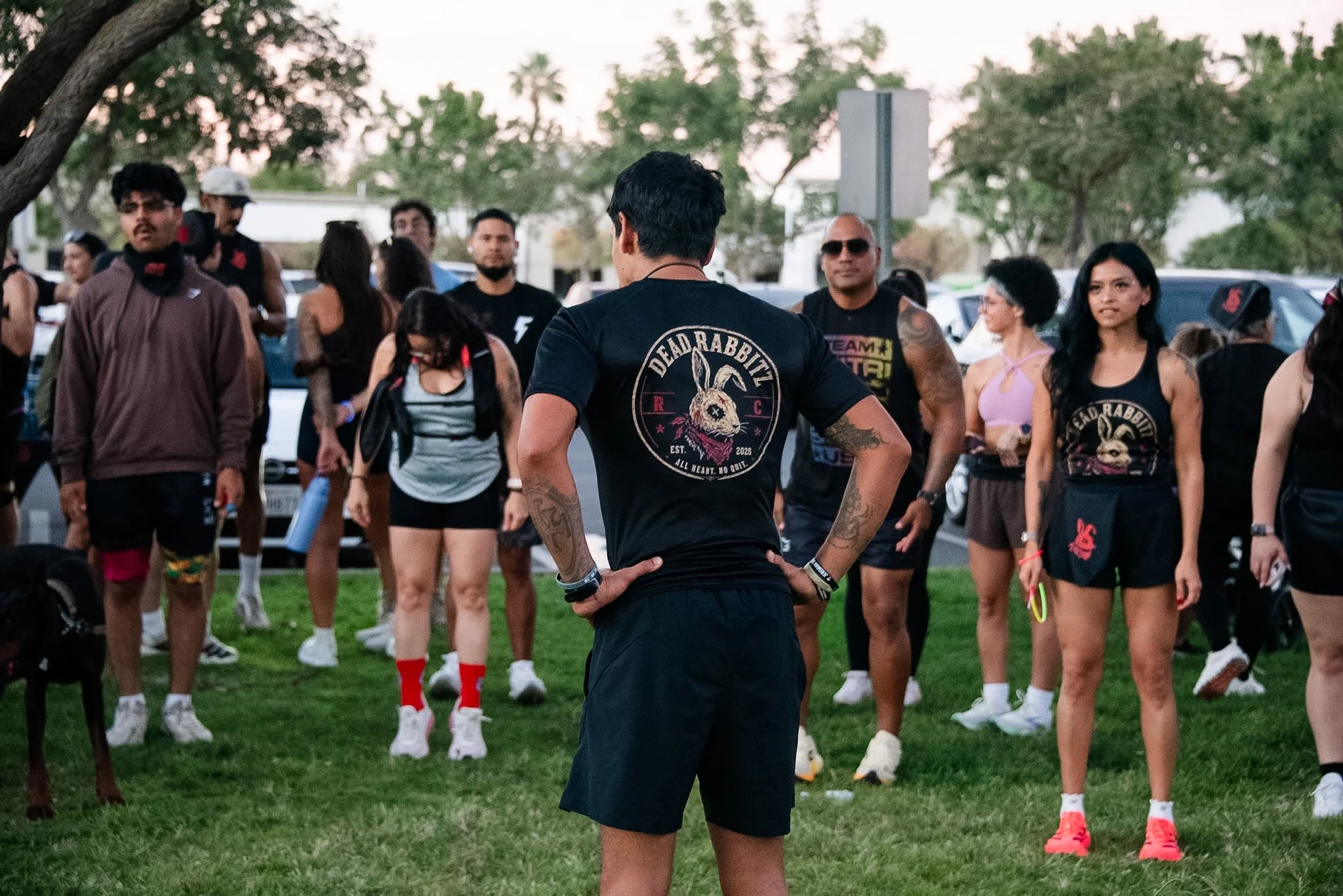 A group of young adults gathered outdoors in a park during daylight, some wearing casual clothes and athletic gear, standing on grass with trees and parked cars in the background. One person in the foreground has their back to the camera, wearing a T