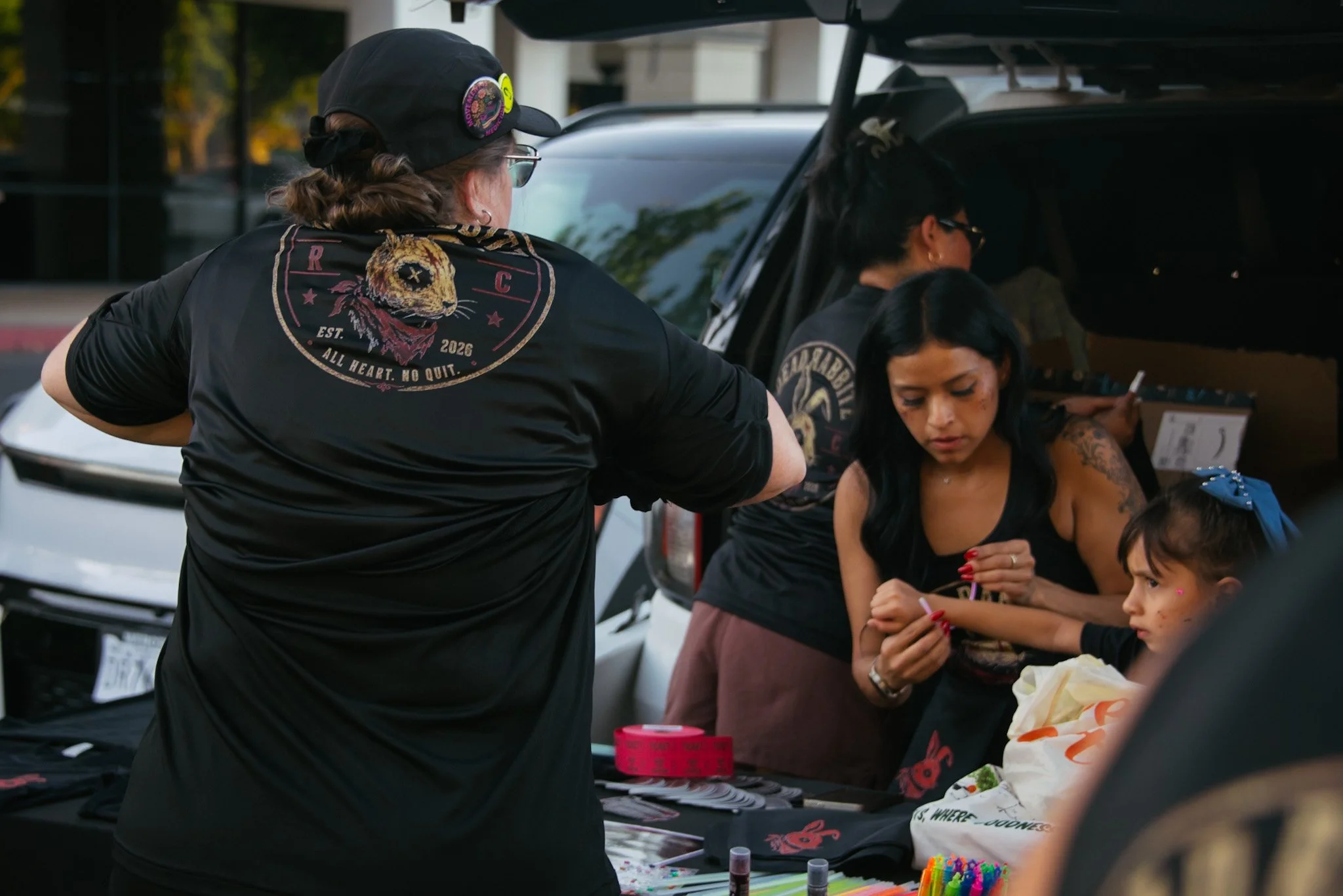 Two women and two children at a table with craft supplies, outdoors near parked cars.
