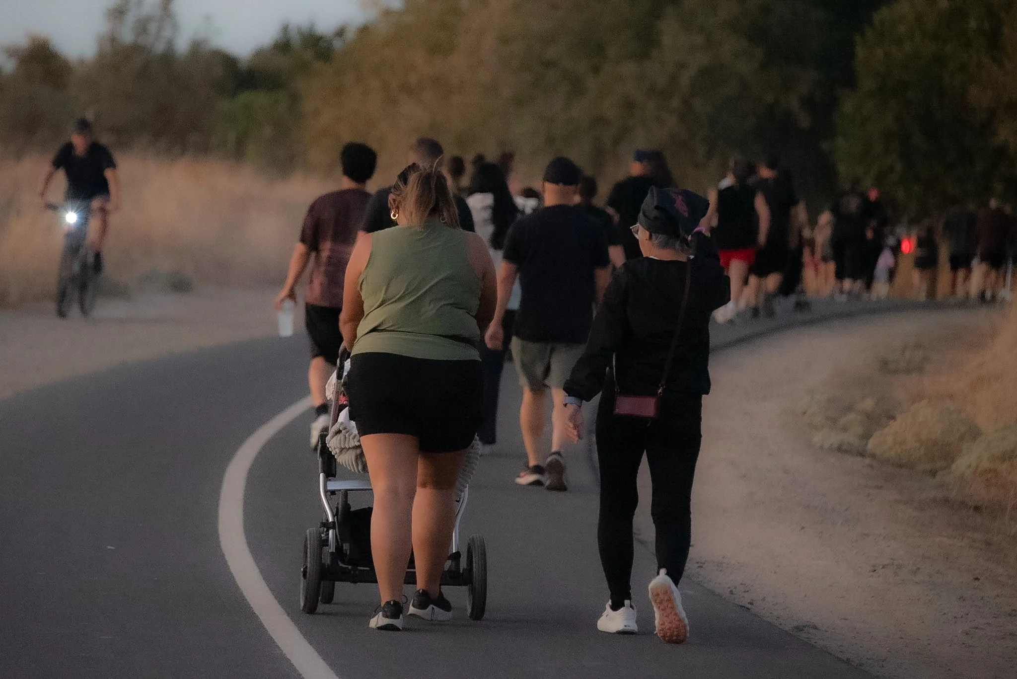 People walking and cycling on a winding road at dusk, surrounded by trees.