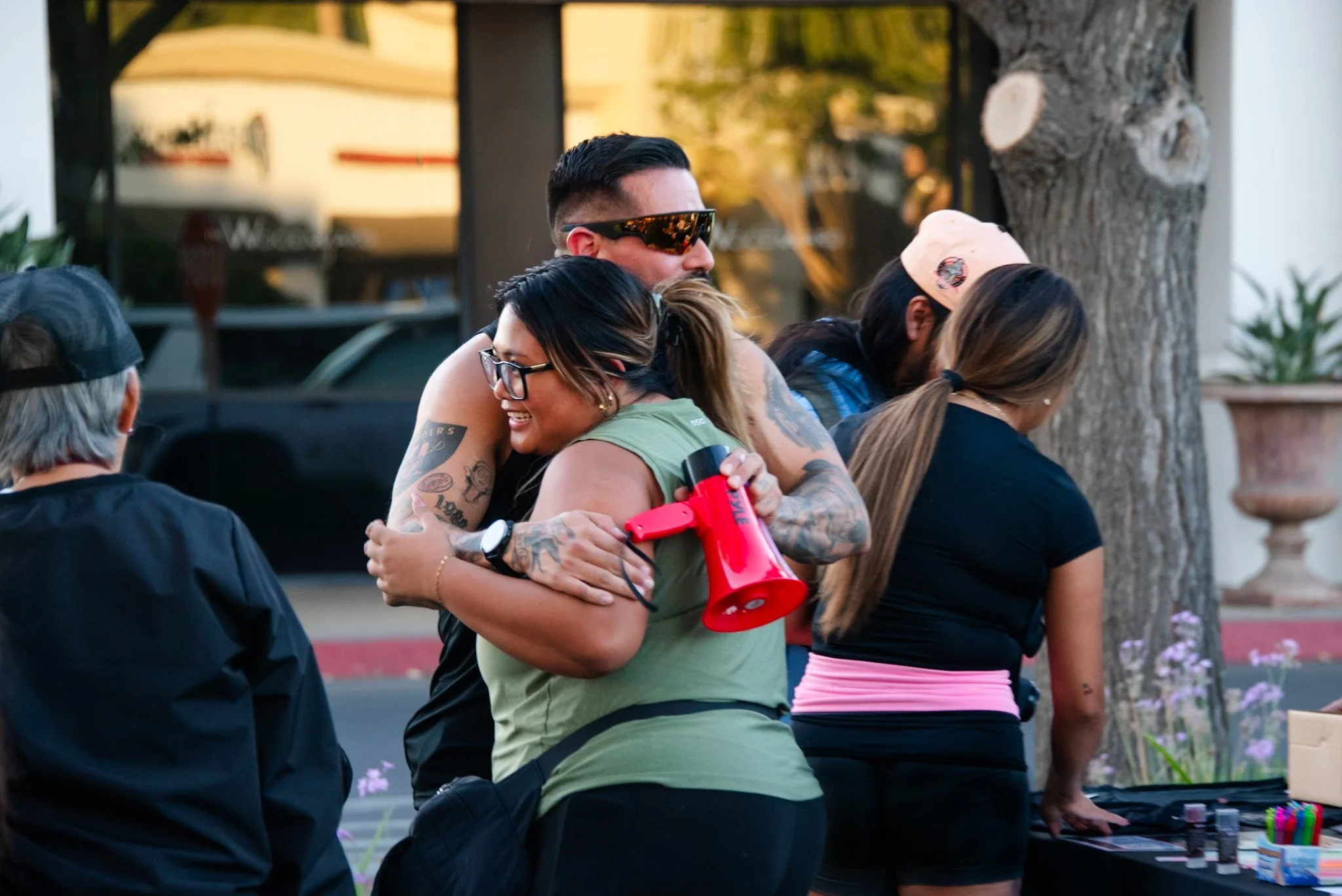 A group of people standing in a line outdoors during the evening, with one person hugging another person from behind. The scene is blurred but appears to be a casual gathering.