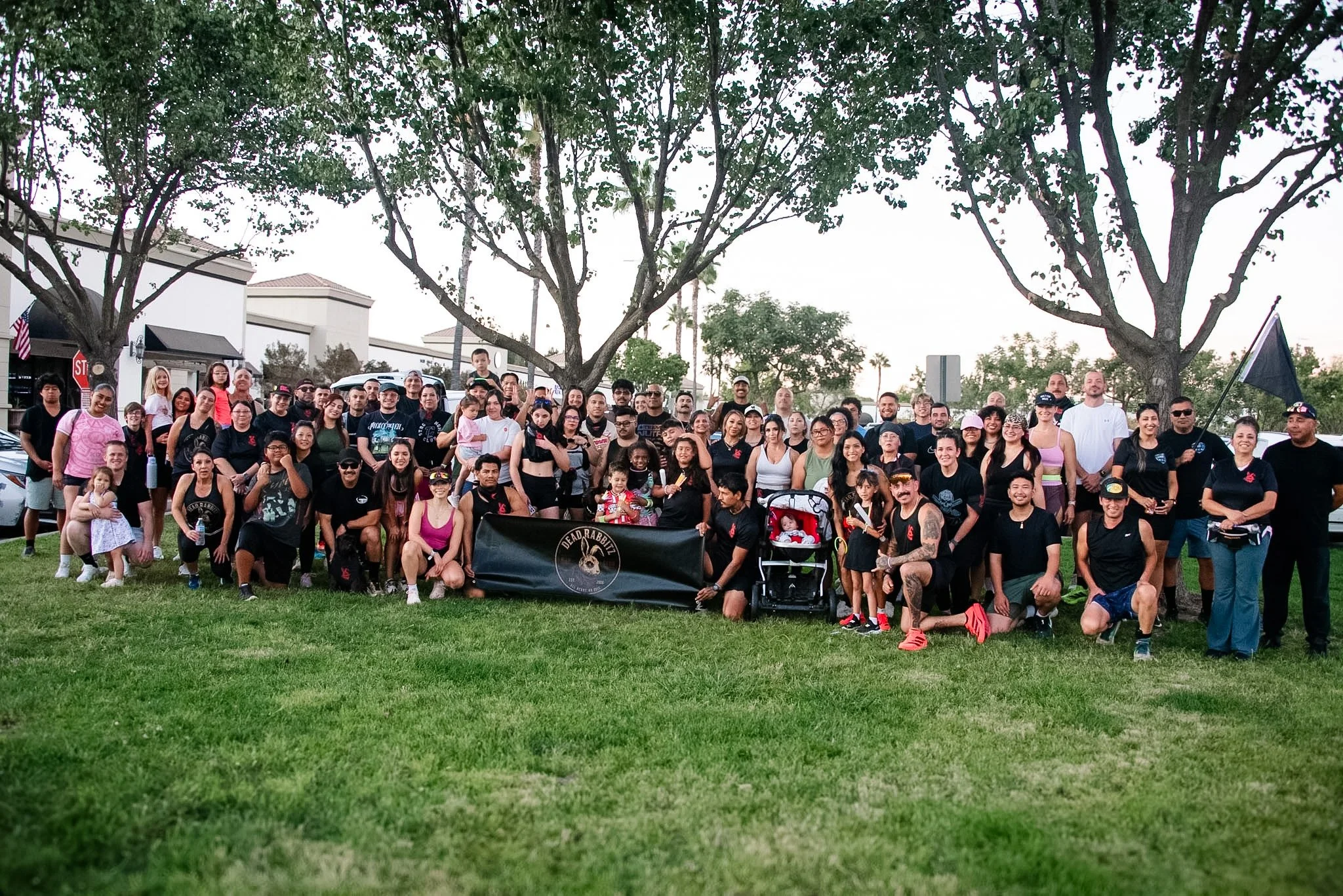 A large group of people gathered outdoors, standing on grass with trees in the background, posing for a group photo on a cloudy day.