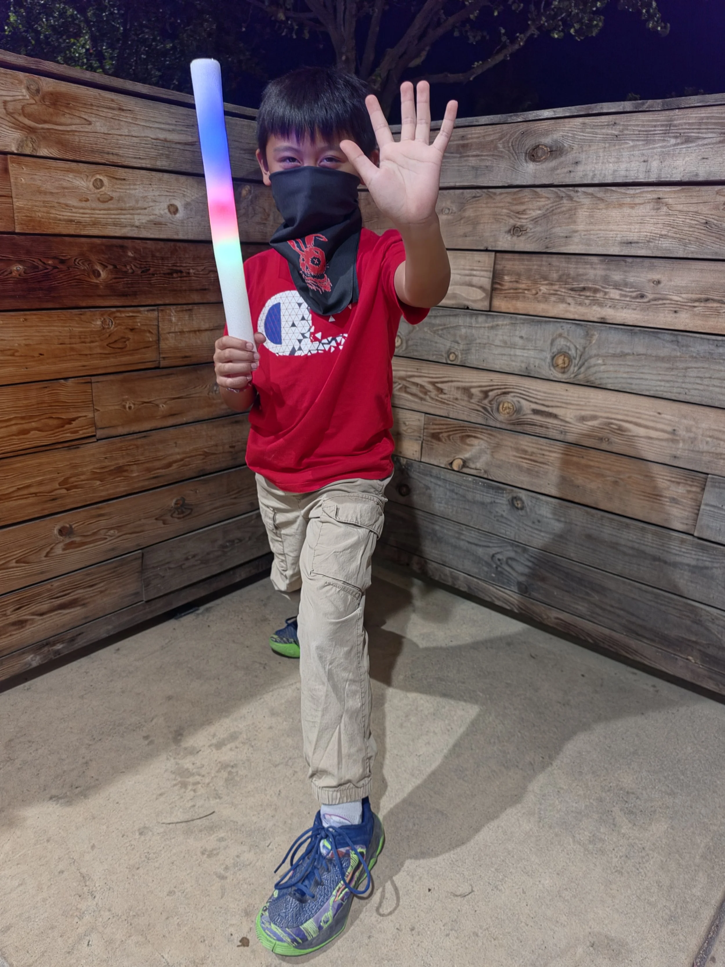 A young boy wearing a black face mask, red t-shirt with a white and blue logo, beige cargo pants, and colorful sneakers, waving and standing on a concrete ground with a wooden fence and dark sky in the background at night, holding a multicolored LED 