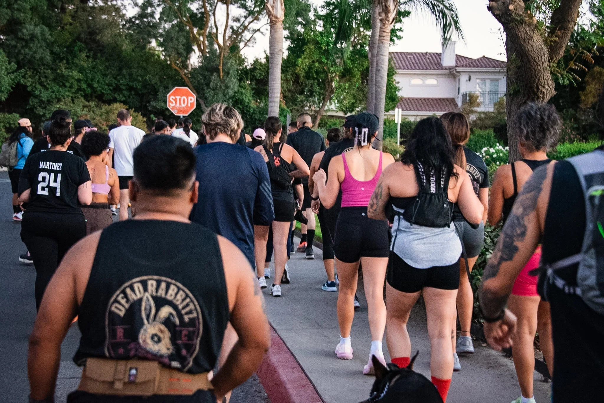 A large group of people participating in an outdoor walk or run event on a residential street with trees, houses, and a stop sign in the background.