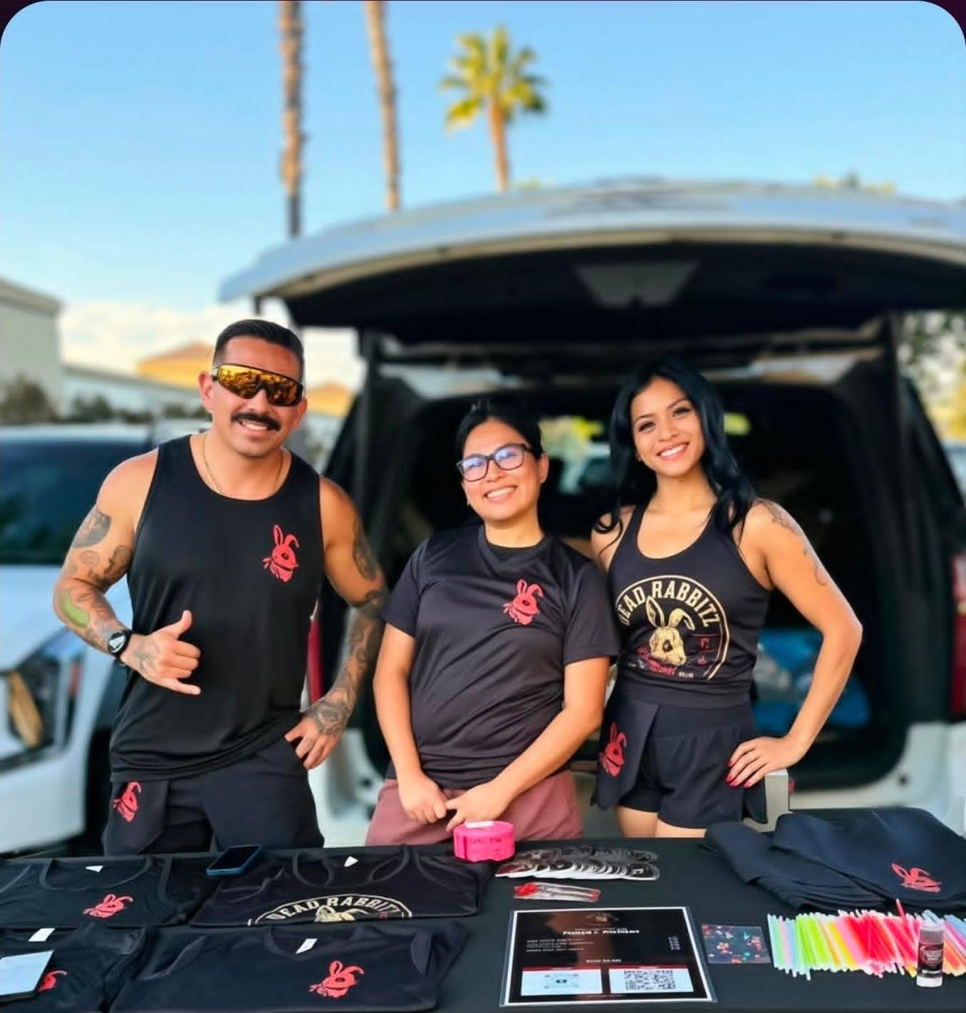 Three smiling people standing in front of an open vehicle trunk at an outdoor event, wearing black T-shirts with a pink rabbit logo, with merchandise and flyers displayed on a table in front of them.