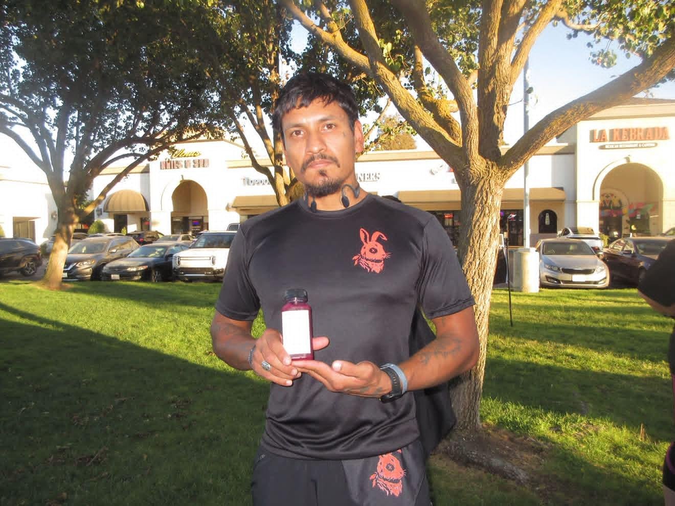 A man standing outdoors in front of trees and a shopping center, holding a small bottle with a white label, wearing a black t-shirt with a red graphic of a rabbit and black shorts.