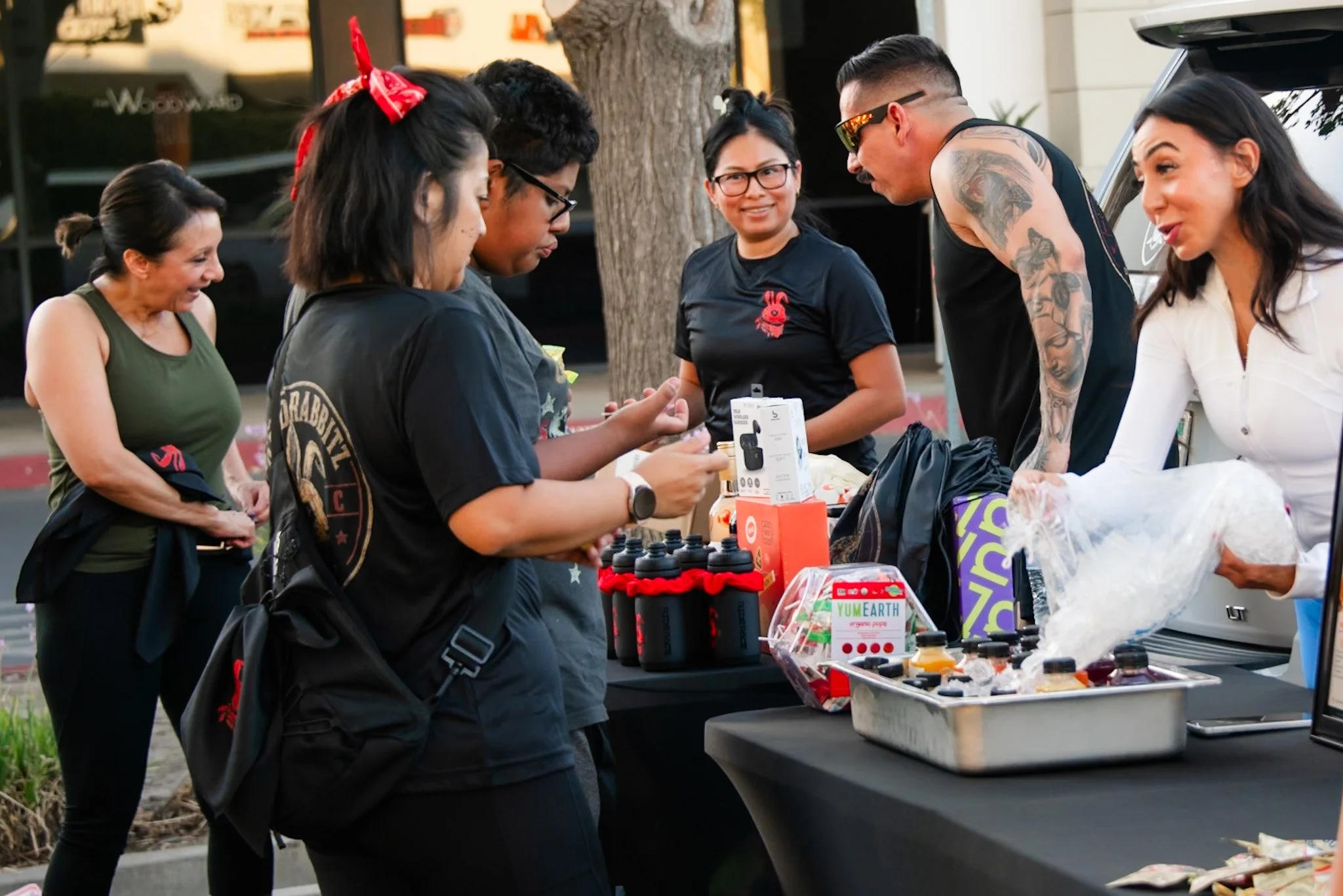 People gathered around a table outdoors, purchasing items at a street market. There are various products, including bottles and packaged goods, on the table.