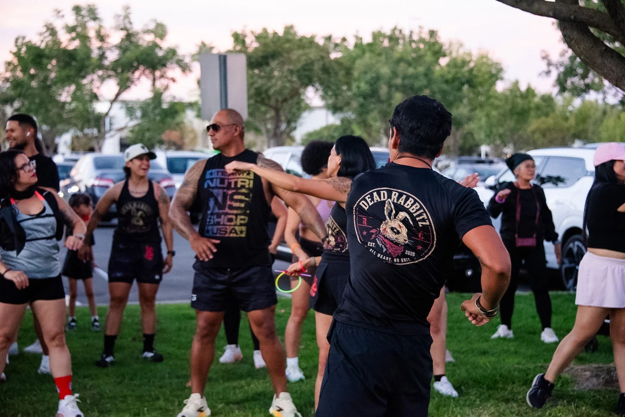 A group of people gathered outdoors on a grassy area, engaging in conversation. Some are wearing athletic clothing and accessories like jump ropes, indicating a fitness or running event. There are cars parked in the background, and trees and a cloudy