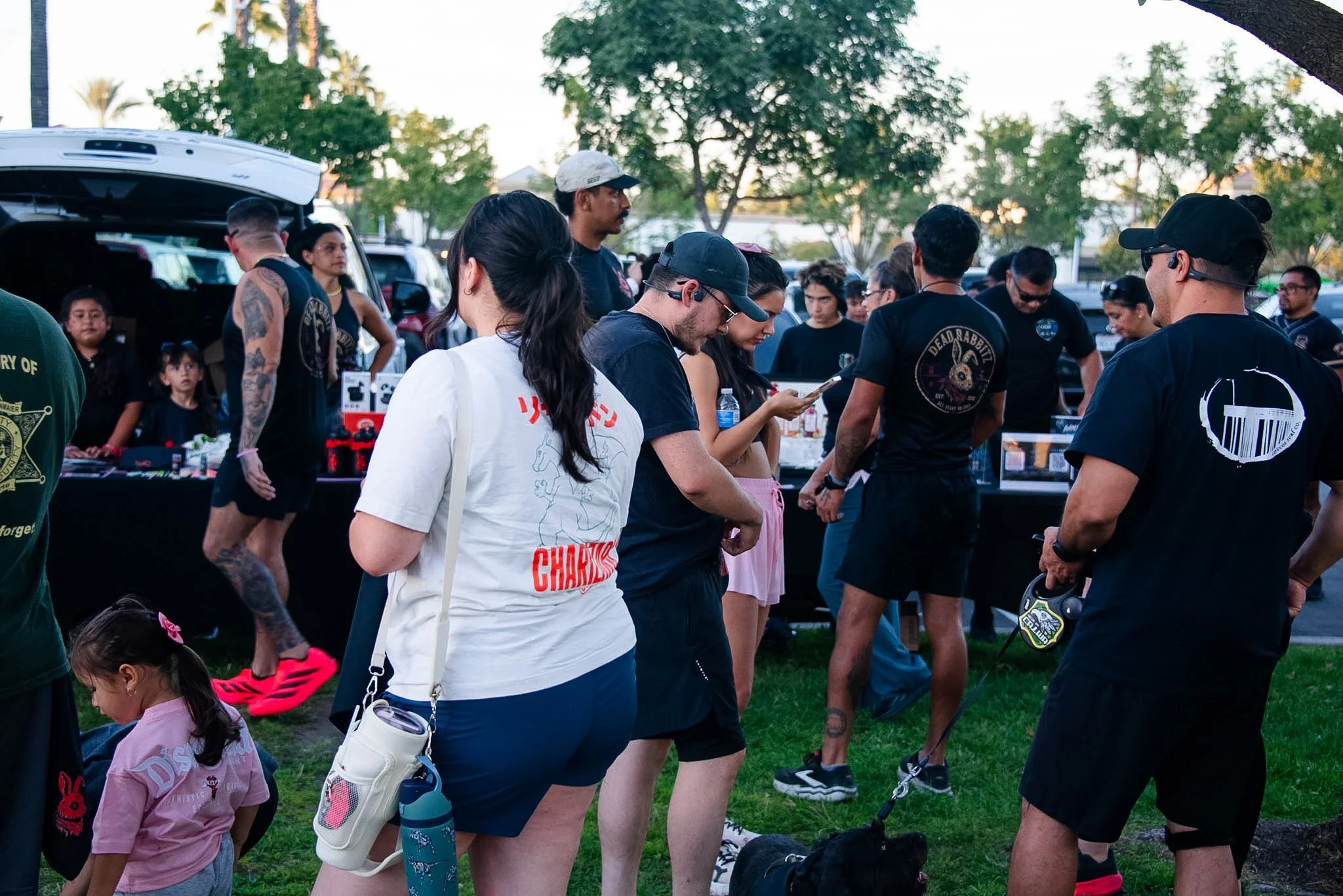 People in a park at sunset, some wearing casual clothes and others in black shirts with logos, gathered near parked cars under trees.