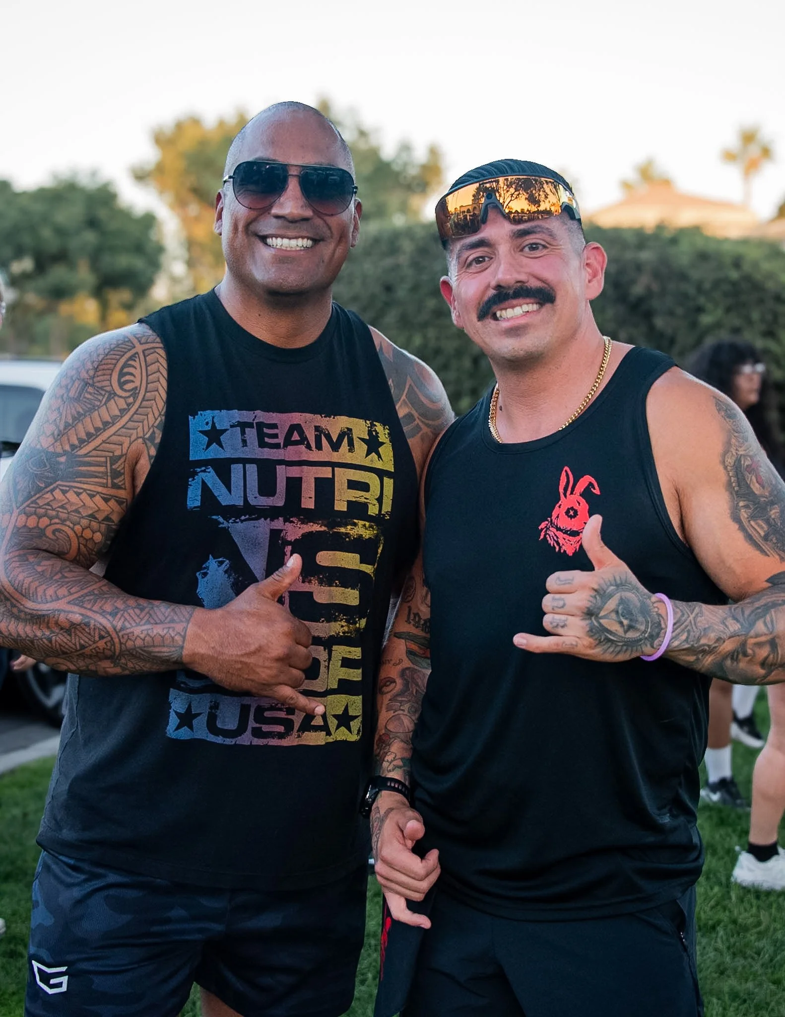 Two smiling men with tattoos on their arms, wearing sunglasses and sleeveless shirts, standing outdoors on grass during daytime.