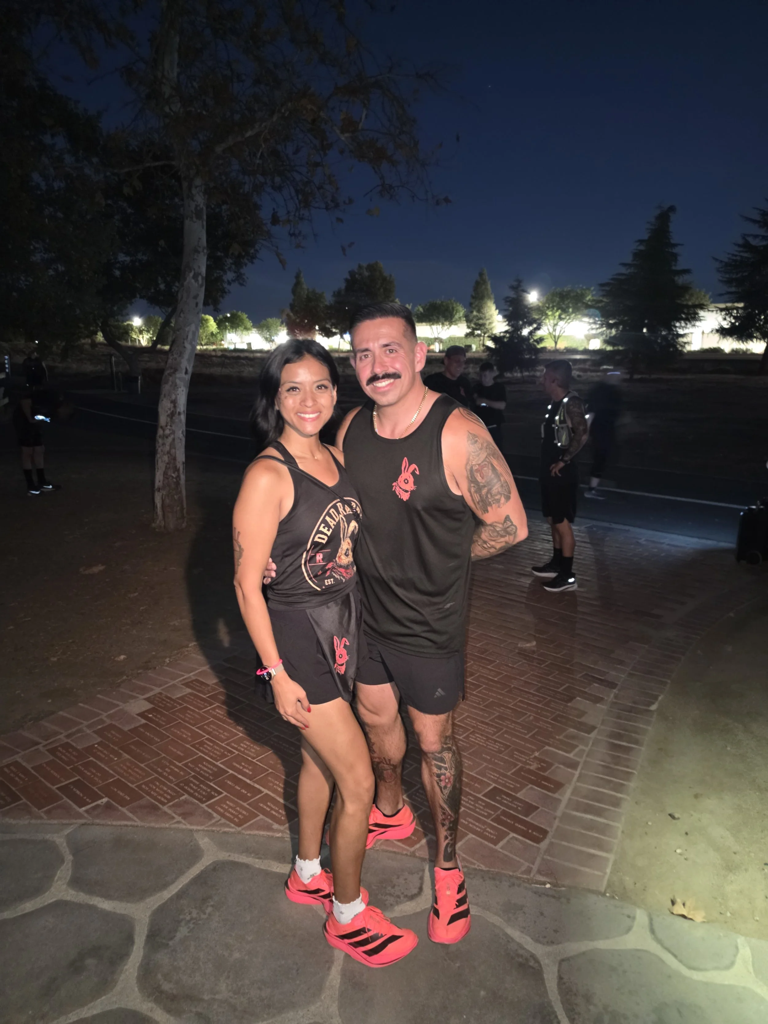 A smiling man and woman are standing close together at night outdoors, dressed in athletic clothing, with a dark sky and trees in the background.