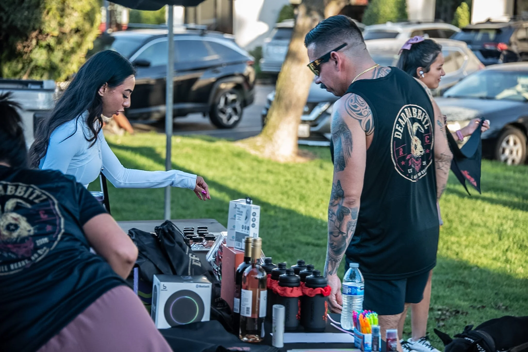 People at an outdoor market or event, with items like water bottles, small boxes, and miscellaneous objects on a table, and cars parked in the background.