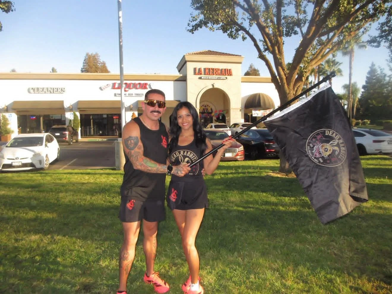 A man and woman standing outside near a parking lot, holding a large black flag with a skull logo, in front of a building called La Kebrada, with cars parked nearby, trees, and a clear sky in the background.