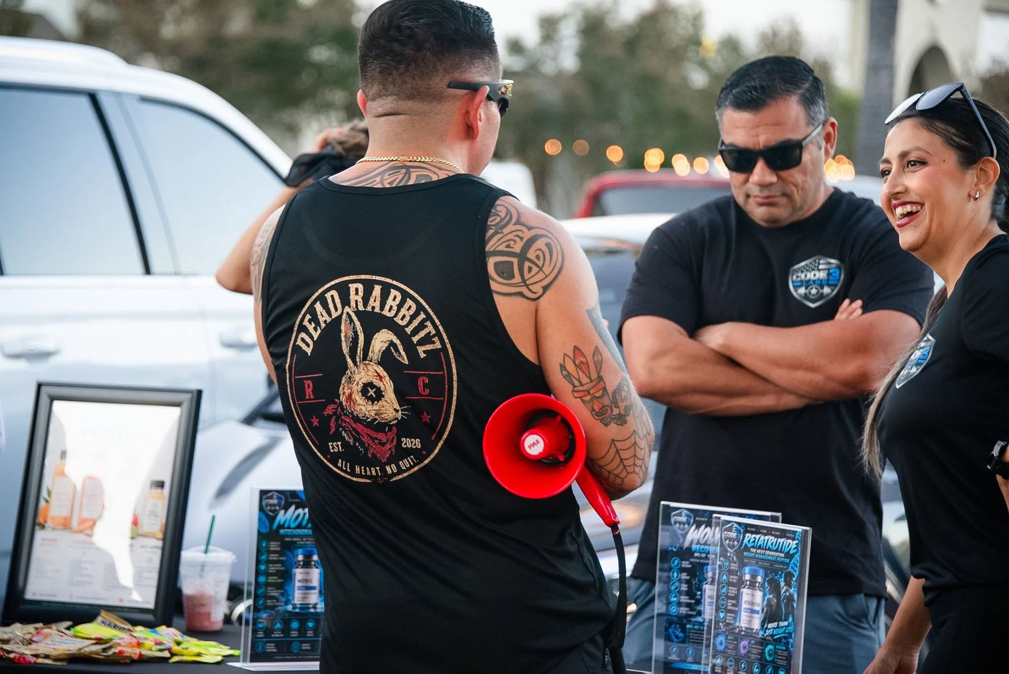 A group of people at an outdoor event, including a man with tattoos wearing a Devil Rabbitz vest, talking to two women, one wearing sunglasses. There are promotional items and informational materials on the table.