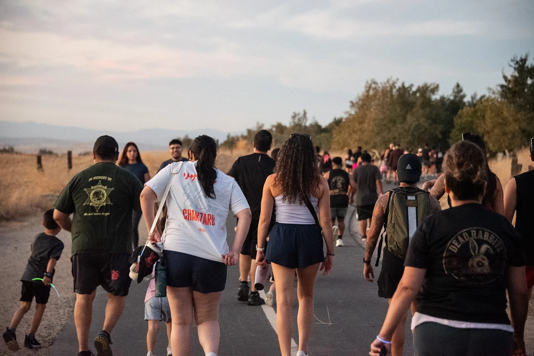 A group of people walking outdoors on a paved path during the early evening, surrounded by trees and open landscape.