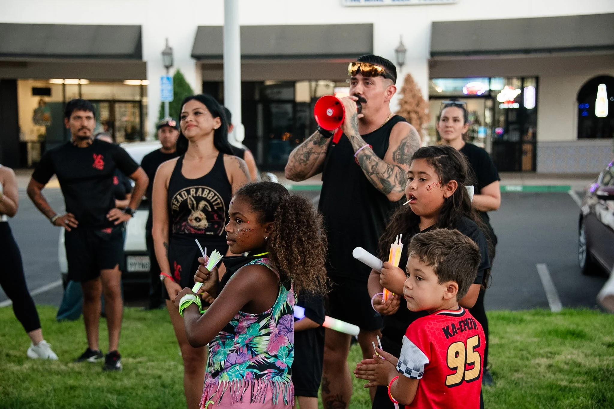 Group of people gathered outdoors, with a man speaking into a red megaphone. Several children and adults are present, some holding glow sticks, and wearing casual attire. It appears to be a community event or gathering.
