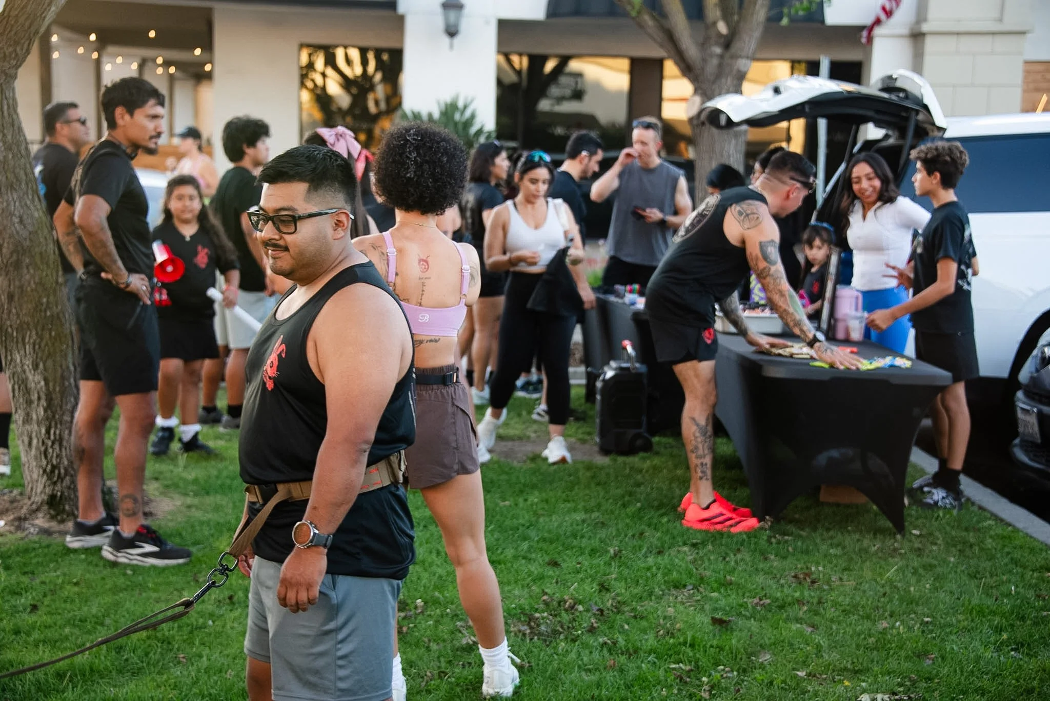 People gathered outdoors at a social event with a table of food, some engaging in conversation, others in line or waiting, with a tree and building in the background.