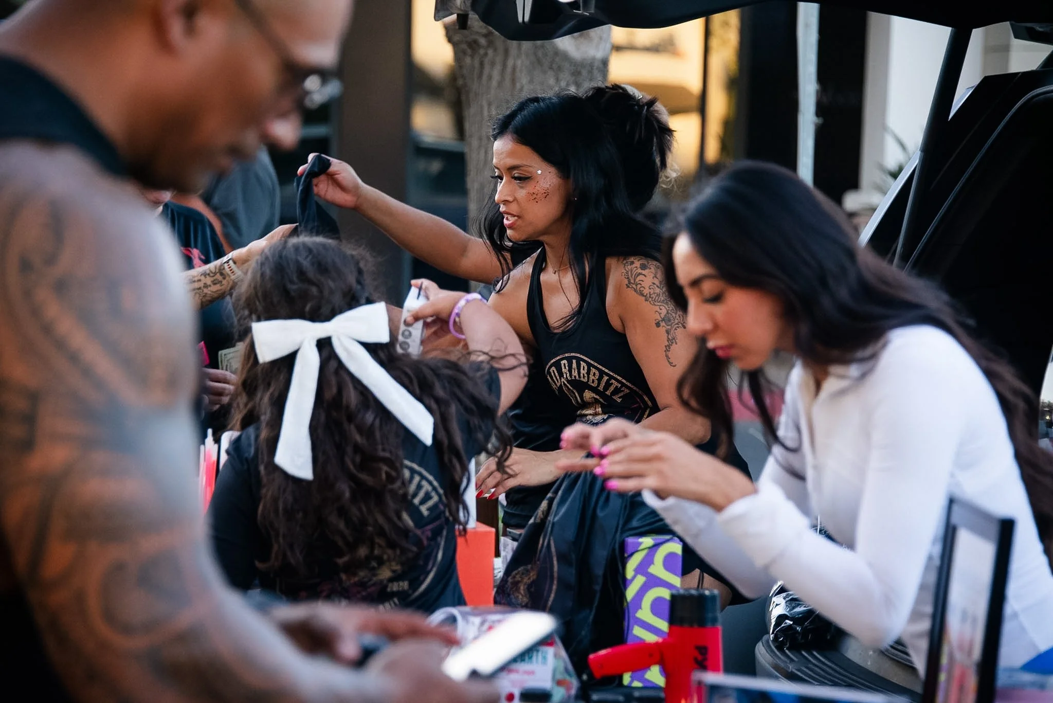 A group of children and adults gathered around a table, engaging in face painting at an outdoor event during the day. The woman in the center has black hair, tattoos on her arm, and is wearing a black tank top, applying face paint to a child's face. 