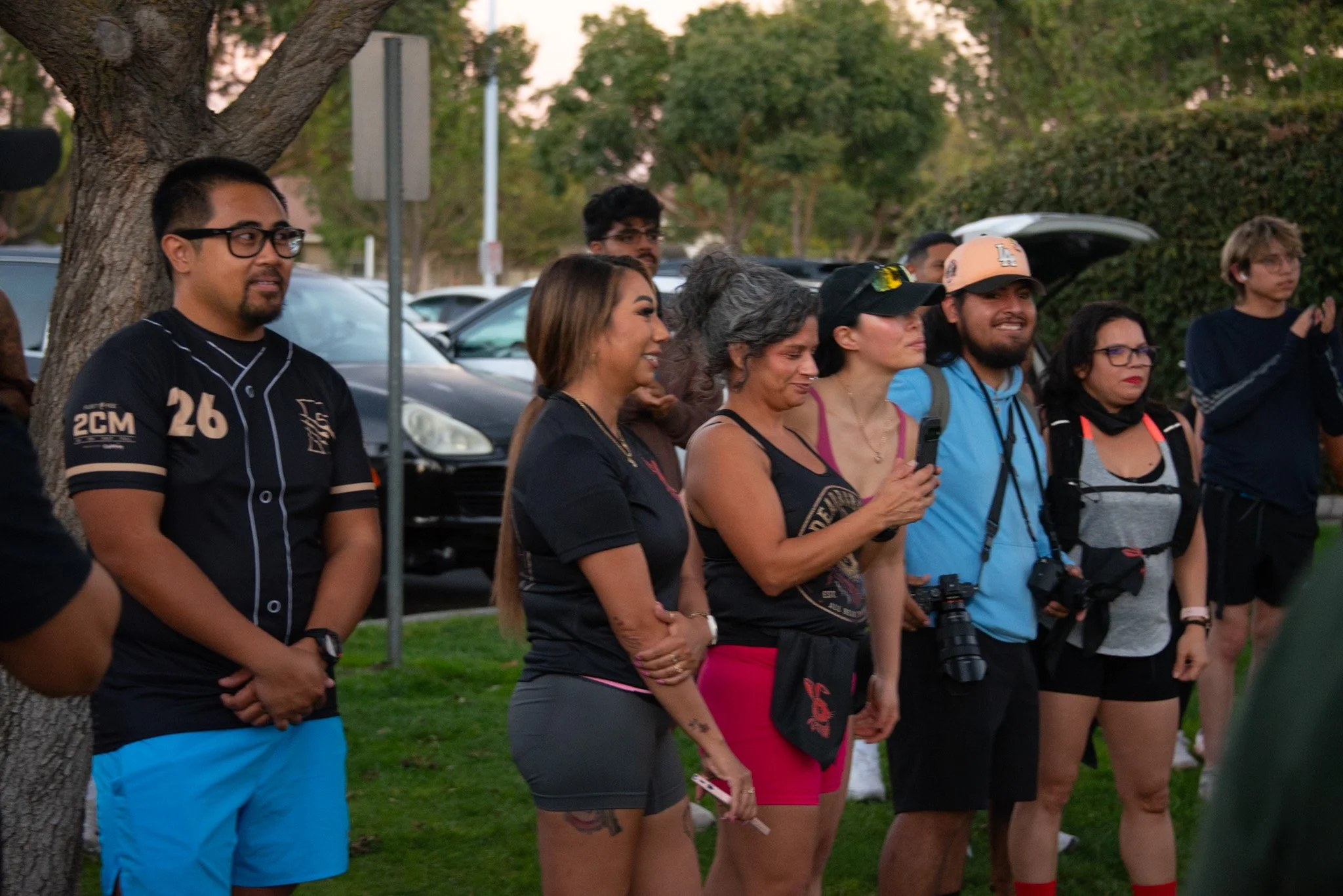 A group of diverse people standing outdoors in front of parked cars, some smiling, some holding cameras, during sunset or early evening.