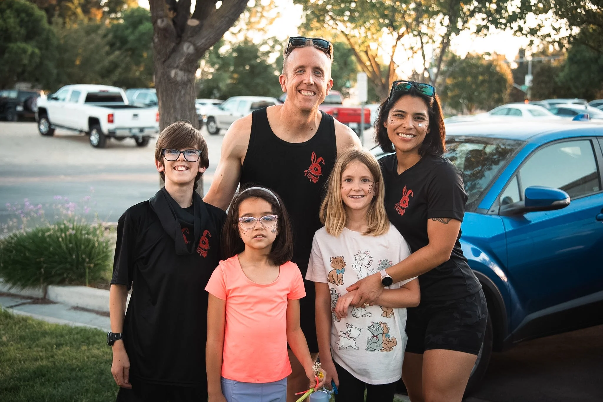 A group of six people, including children and adults, smiling outdoors in a parking lot near trees and cars during sunset.