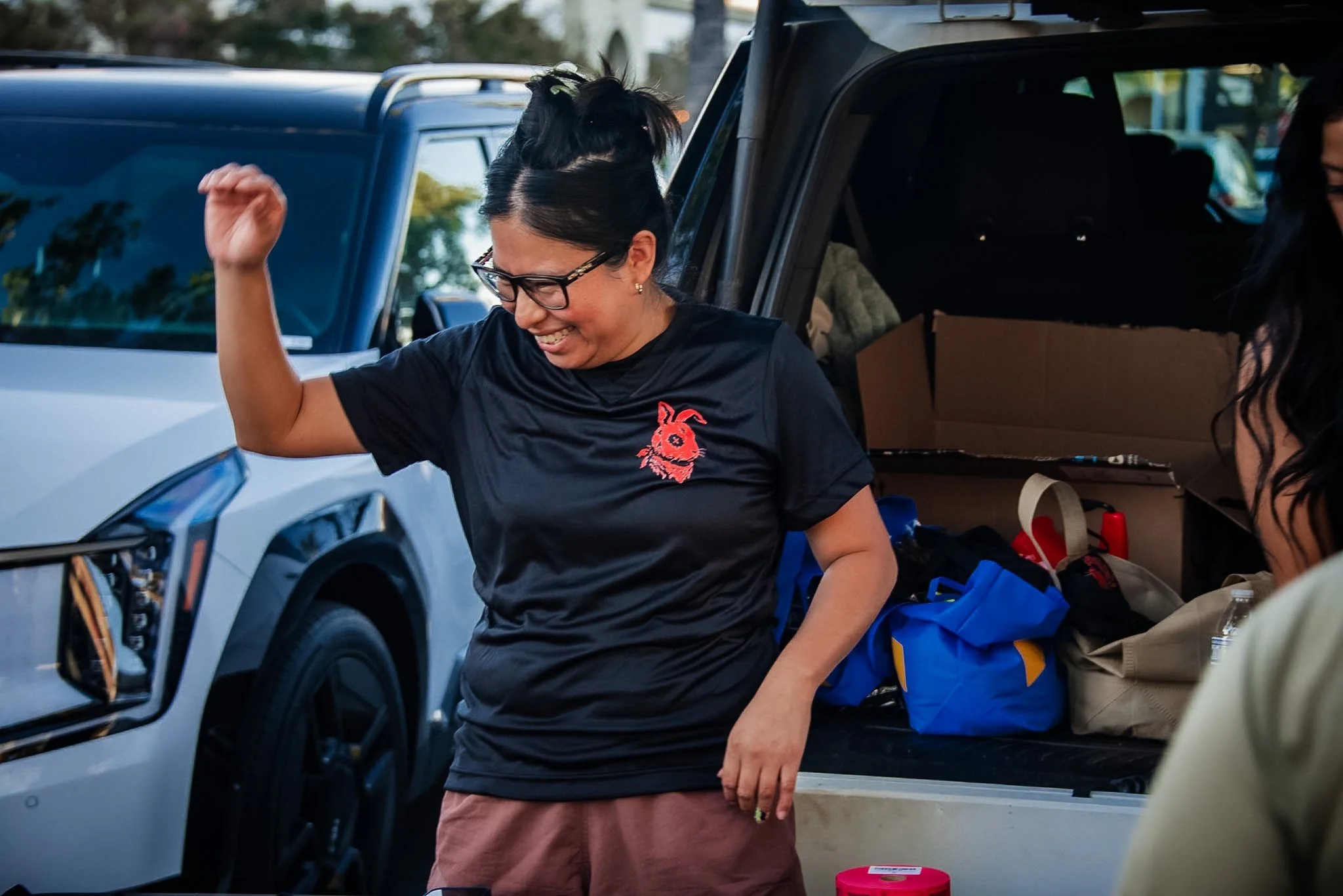A woman with glasses and dark hair tied up in a bun, smiling and raising her arm, standing beside an open car trunk filled with bags and boxes.