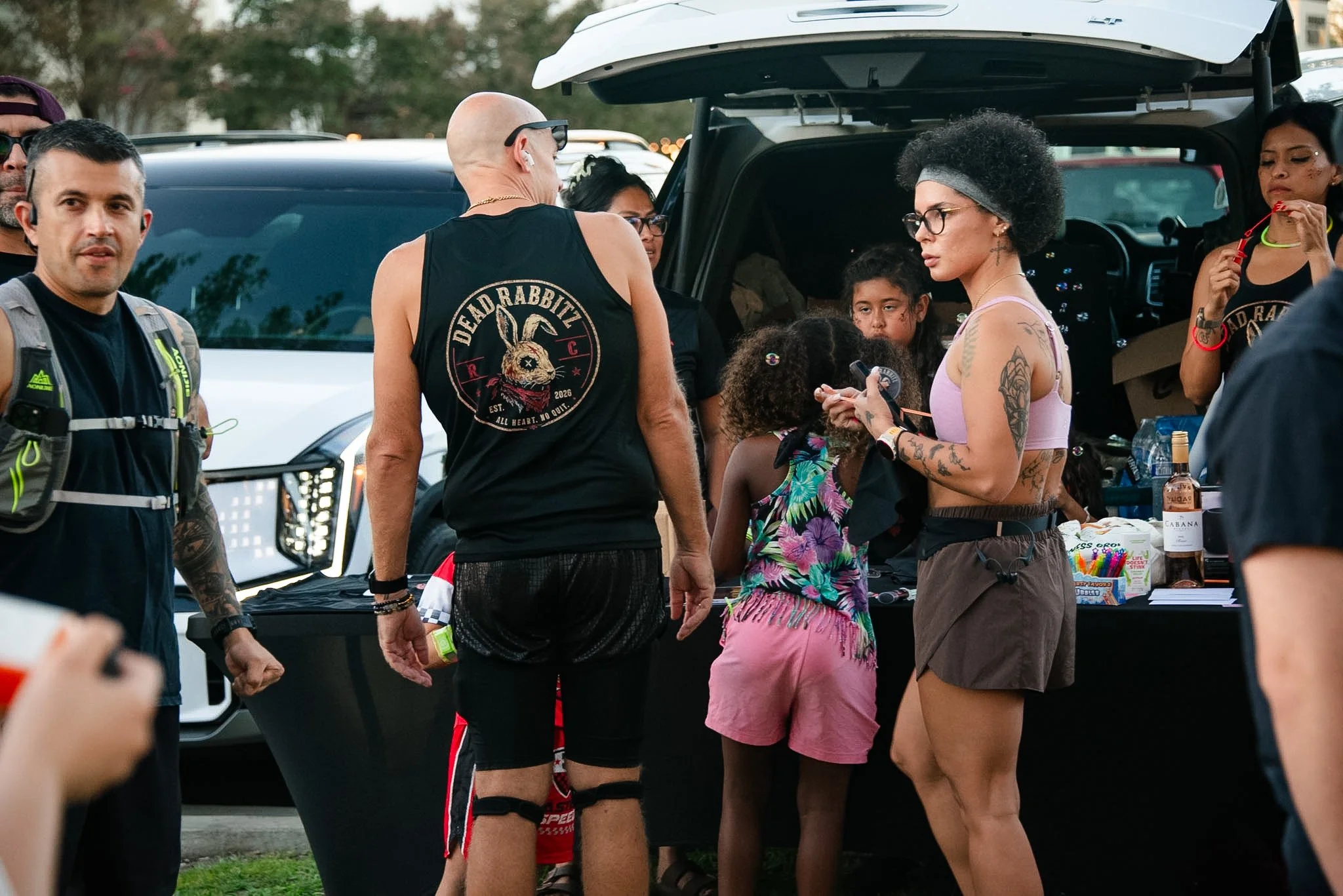 Group of people gathered around a black vehicle, some women wearing tank tops and glasses, a man wearing a sleeveless shirt with a rabbit graphic, and children at an outdoor event with tables loaded with snacks and drinks.