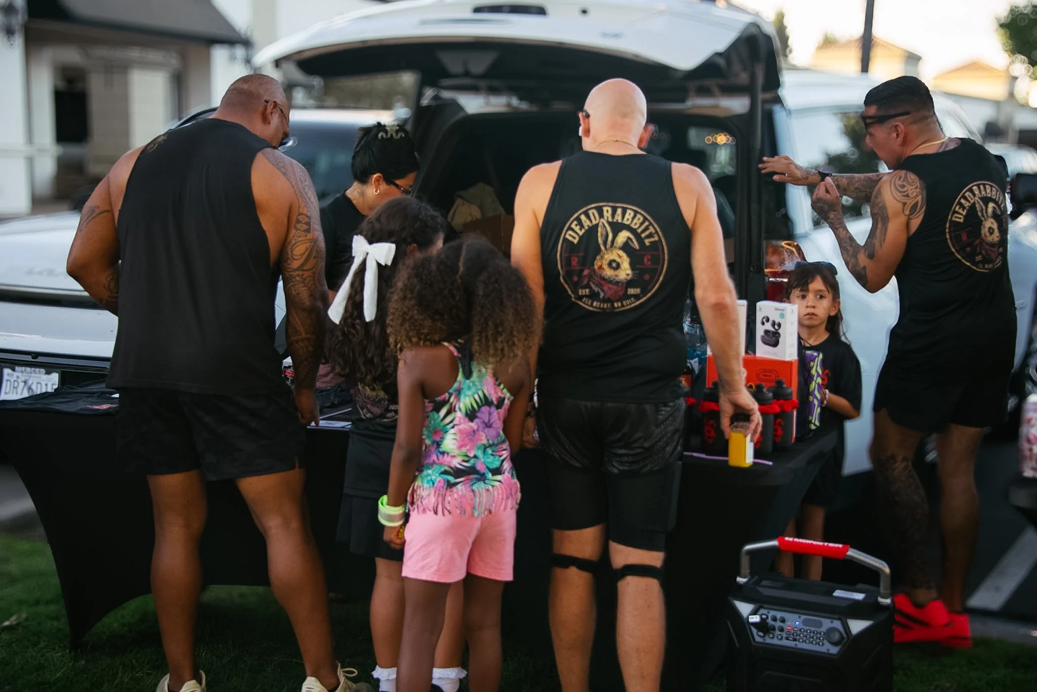 A group of people, including children, gathered around the open trunk of a SUV during a community event. Some individuals wear sleeveless shirts with a logo that reads 'Dead Rabbits.' Children are inspecting items on a table, including a box of wirel