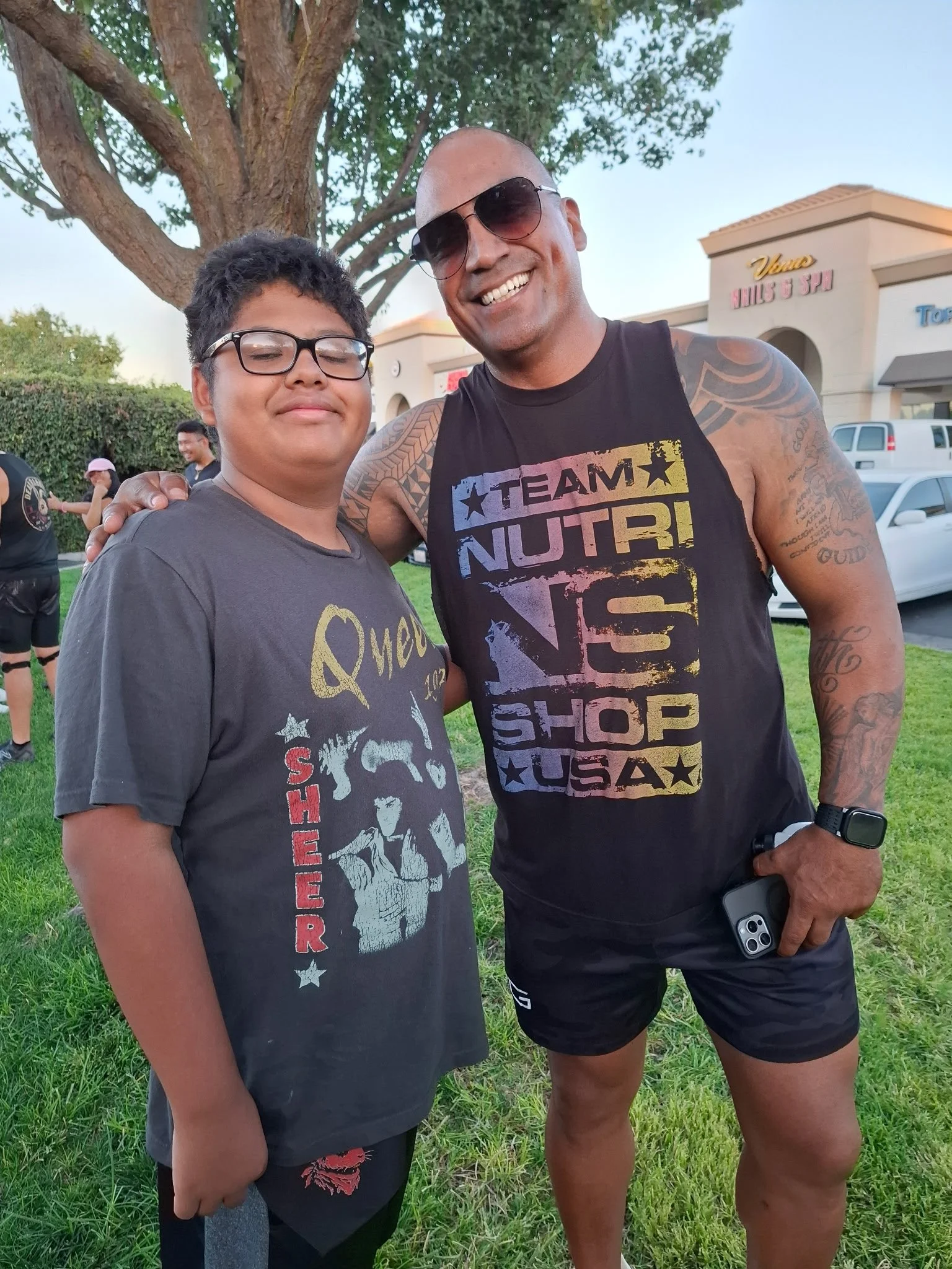 A man and a boy standing outdoors under a tree, smiling at the camera. The man is wearing sunglasses and a sleeveless shirt with text, and the boy is wearing glasses and a T-shirt with graphic print. They are in a grassy area with a shopping plaza in