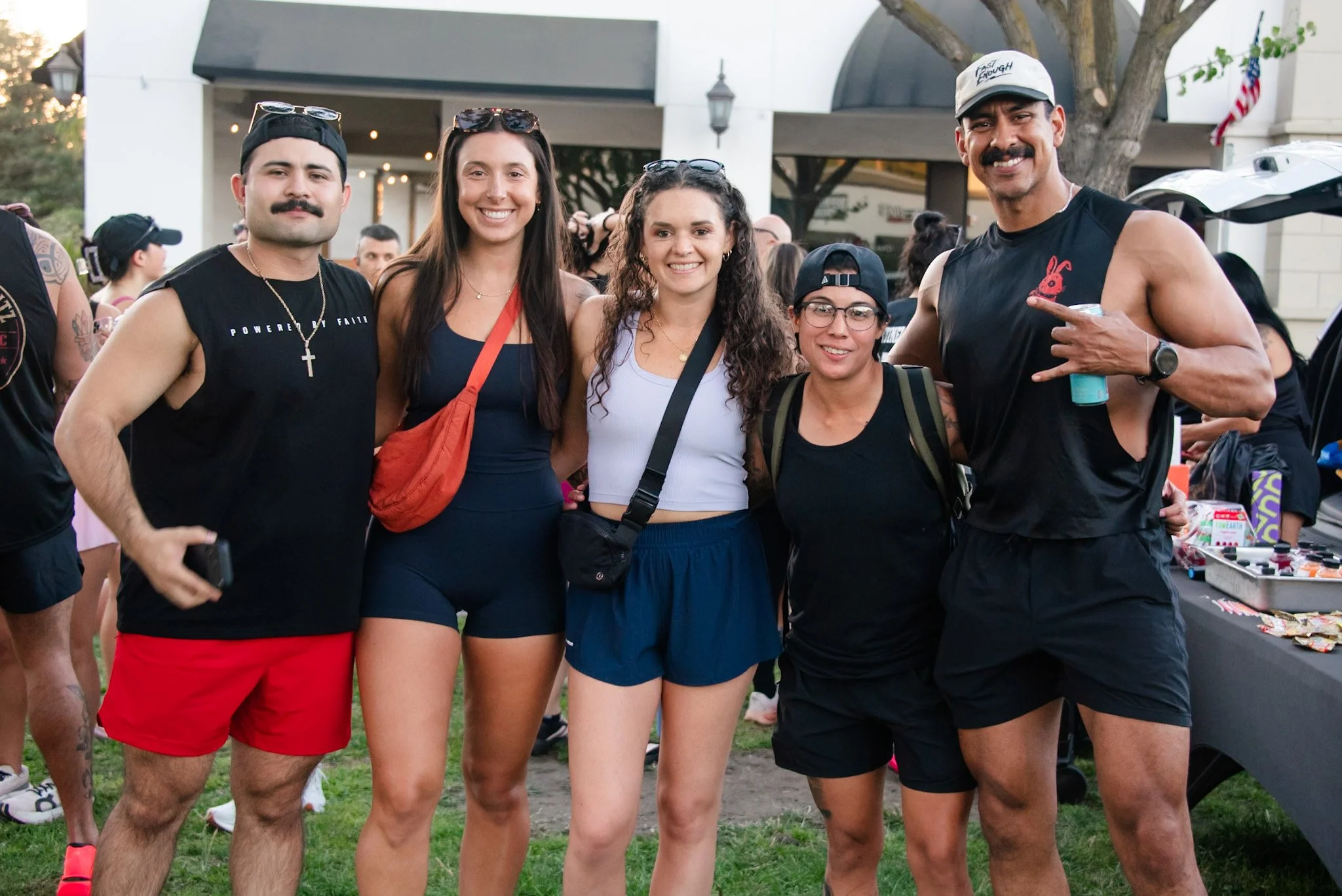 Group of five diverse young adults standing outdoors at an event, smiling and posing for the photo, with a table of snacks and other people in the background.