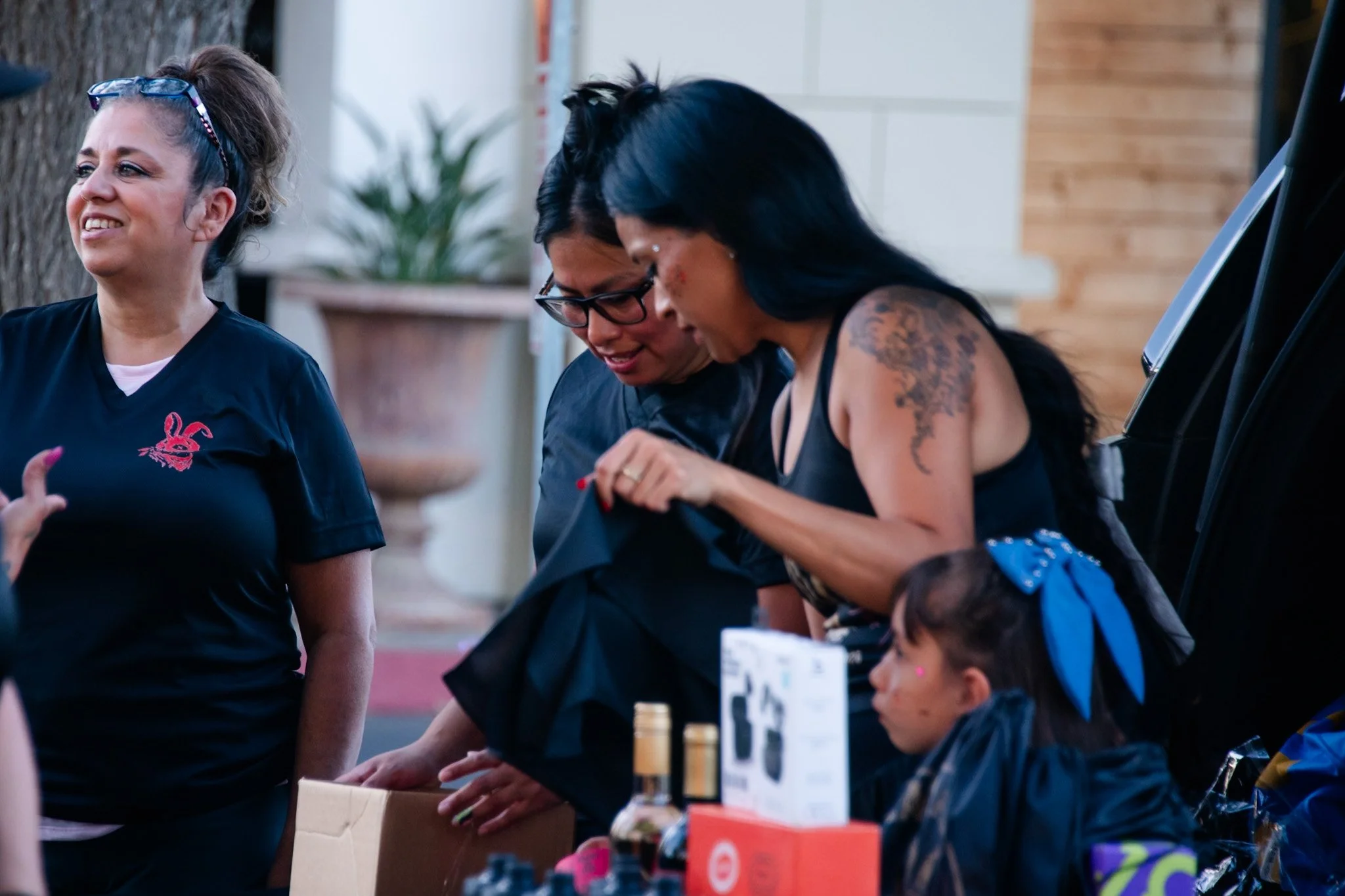 Group of women interacting at a booth or market, with one woman showcasing tattoos, another wearing glasses, and a third woman smiling and wearing sunglasses on her head.