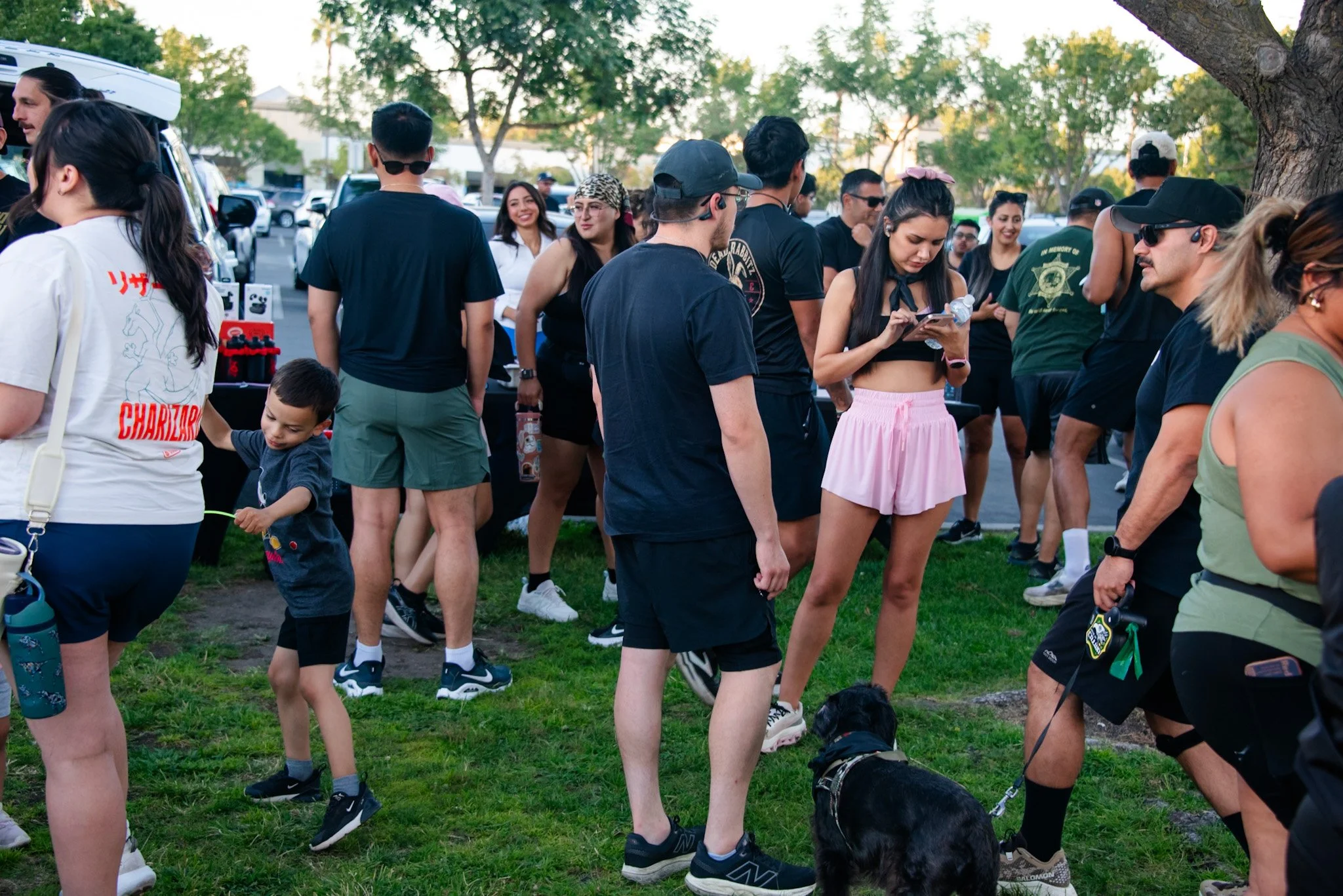 People gathered outdoors in a park, some interacting with police officers and service dogs, with trees and a parking lot in the background.