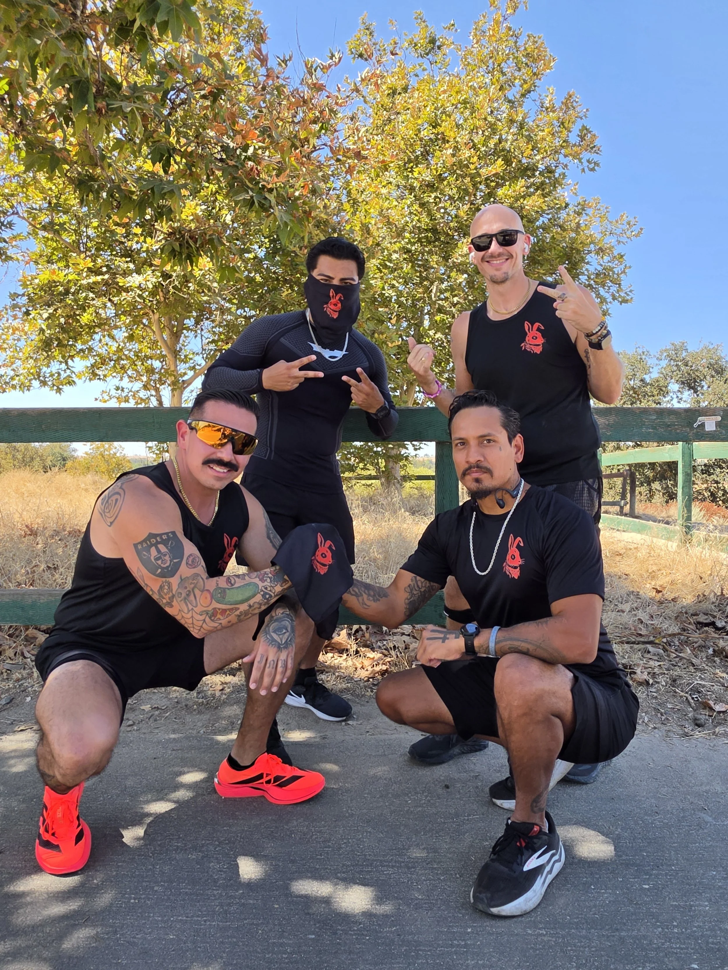 Group of five men outdoors posing for a photo, some squatting and others standing, under a large tree with autumn-colored leaves. They are dressed in casual athletic wear, some with sunglasses and accessories, showing a friendly and energetic vibe.