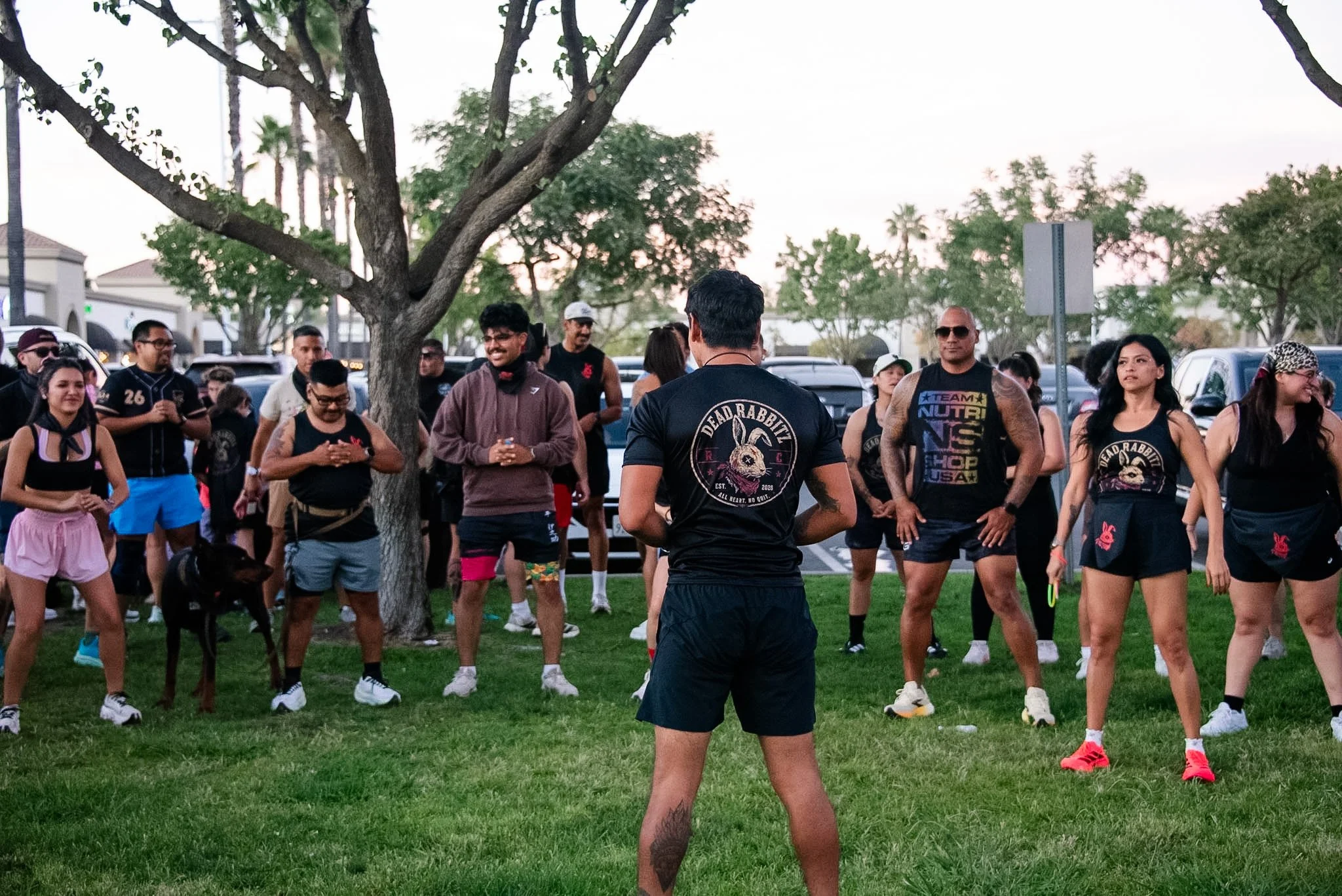 A group of people gathered outdoors in a park, some wearing athletic clothing, with a man in the foreground facing a crowd, possibly addressing or instructing them, near a large tree during sunset.
