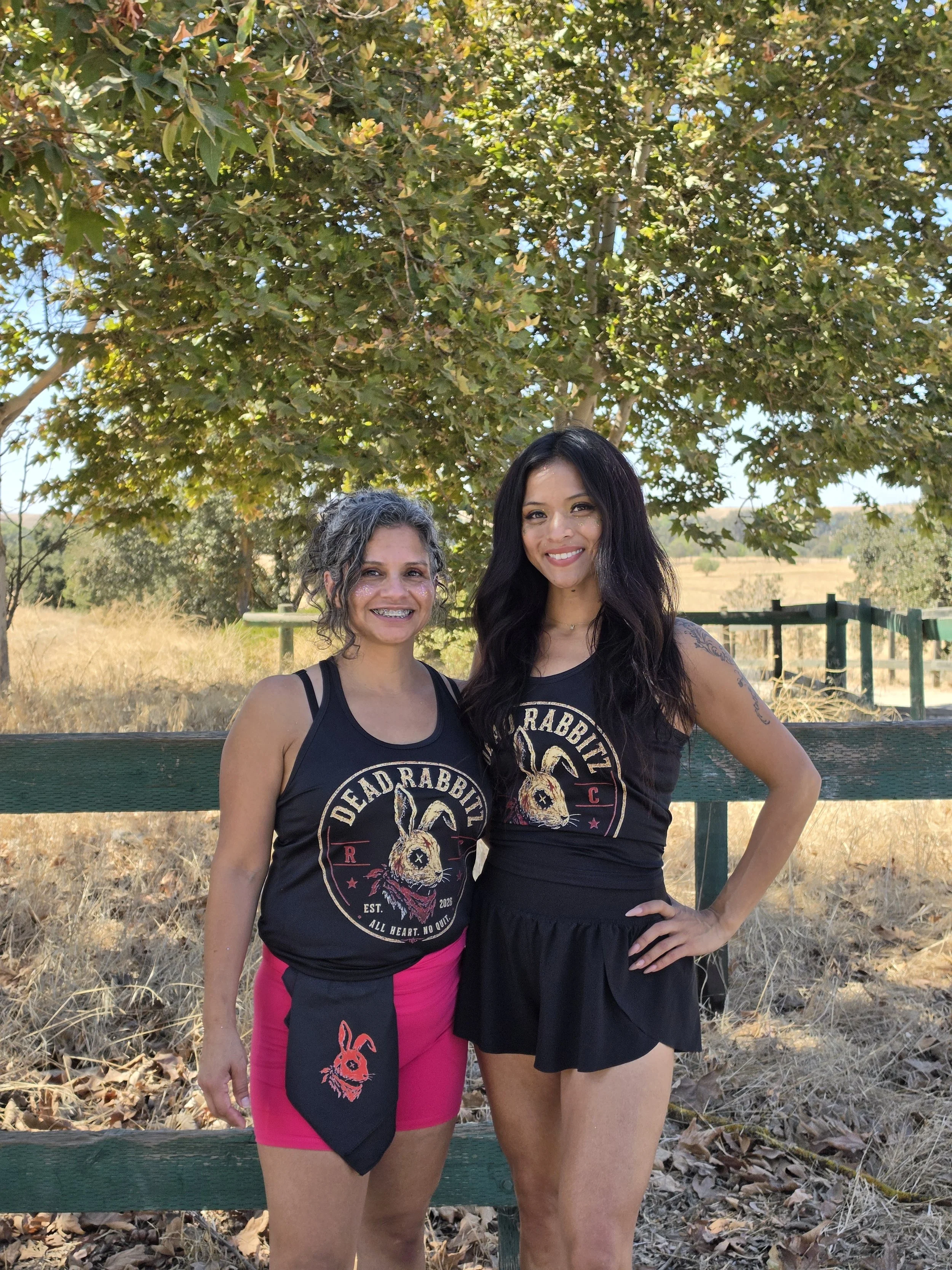 Two women standing outdoors in front of a tree, smiling at the camera. They are wearing black sleeveless t-shirts with a graphic of a rabbit and the words 'Dead Rabbitz'. One woman has curly gray hair and is wearing pink shorts, while the other has long black hair and is wearing a black skirt.
