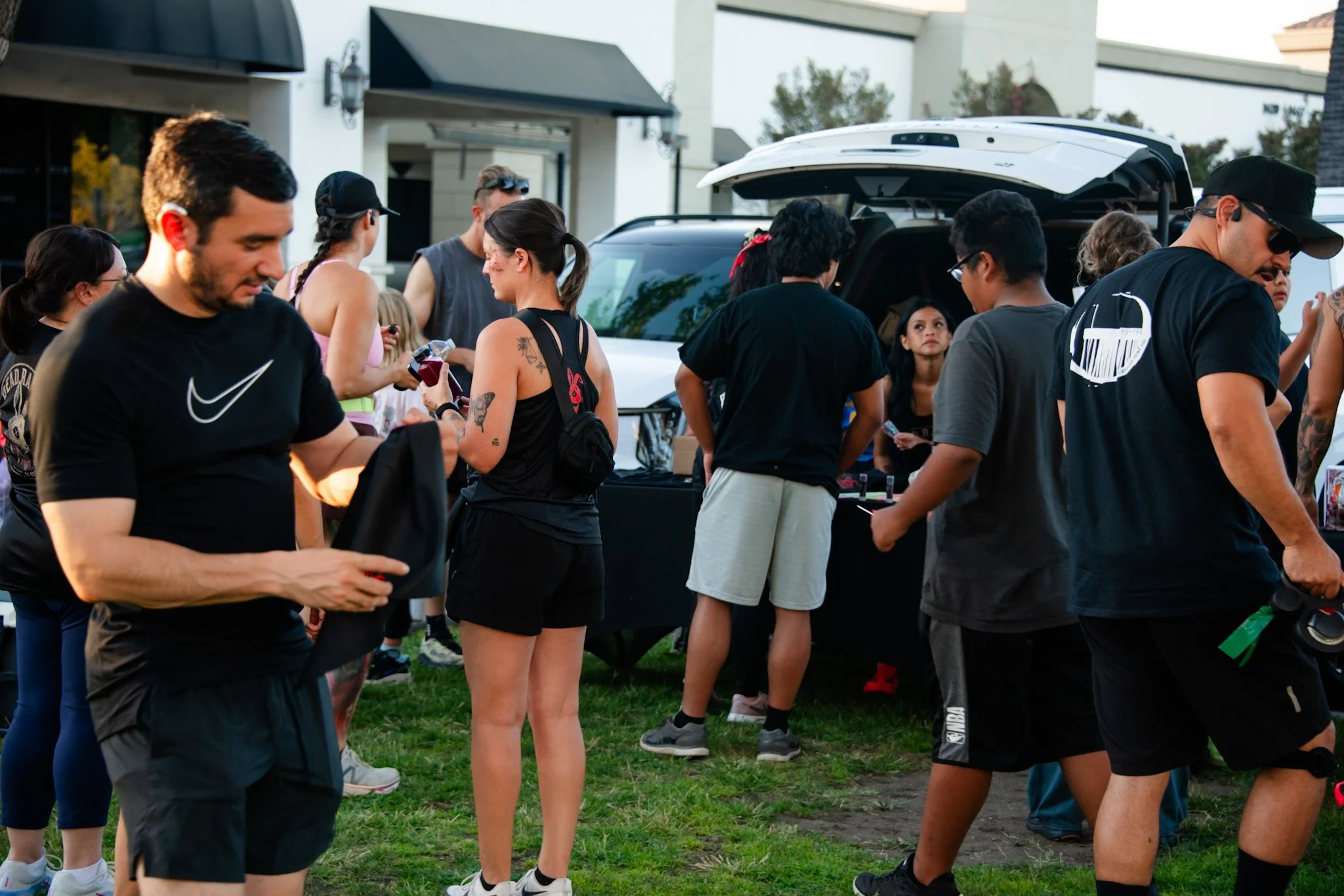 A group of people gathered outdoors near a vehicle with an open trunk, engaging in conversation and activities, with a building and trees in the background.