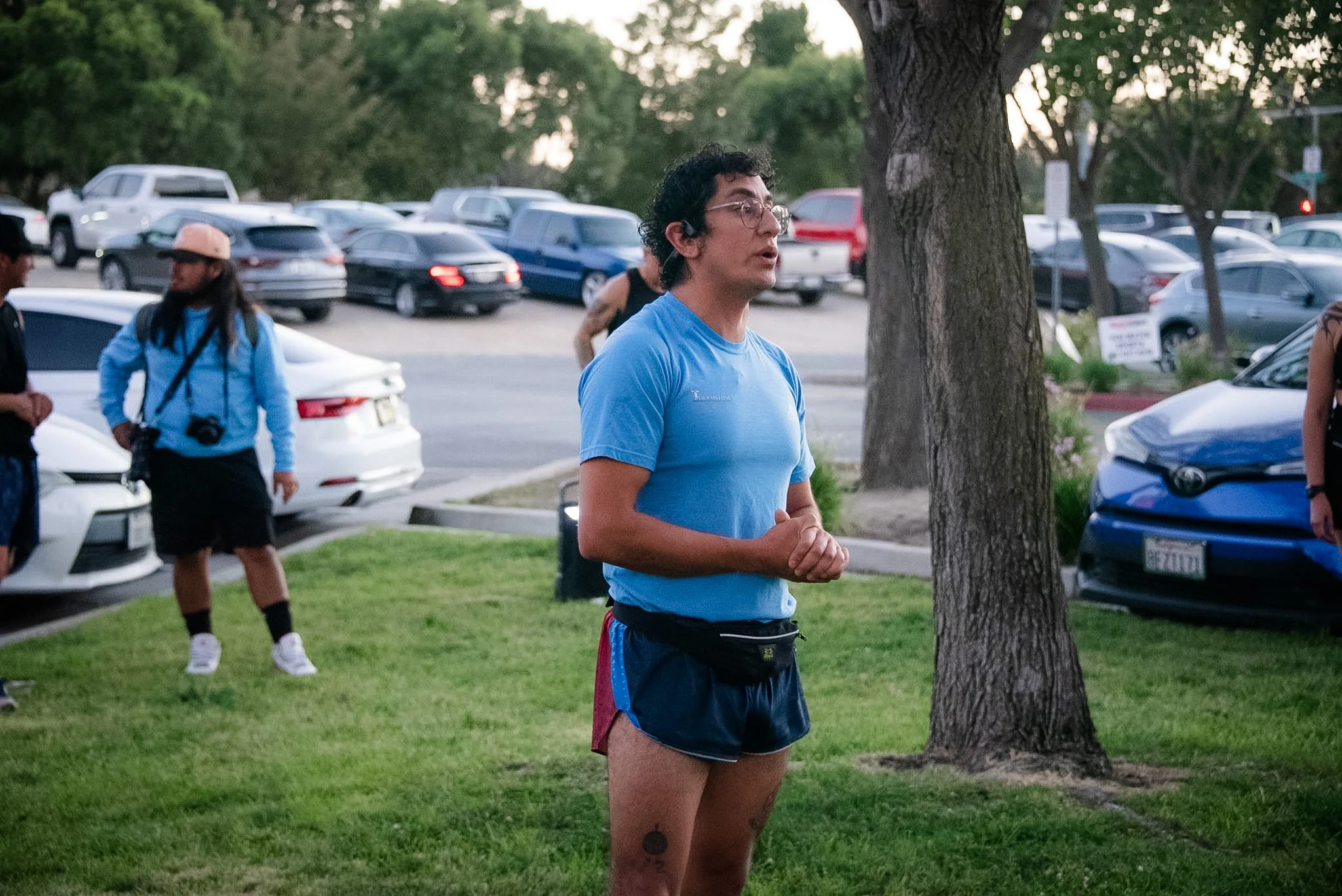 A young man with curly dark hair, glasses, and tattoos, standing outdoors behind a tree, wearing a blue T-shirt and shorts. Several people and parked cars are visible in the background, suggesting a park or gathering area.