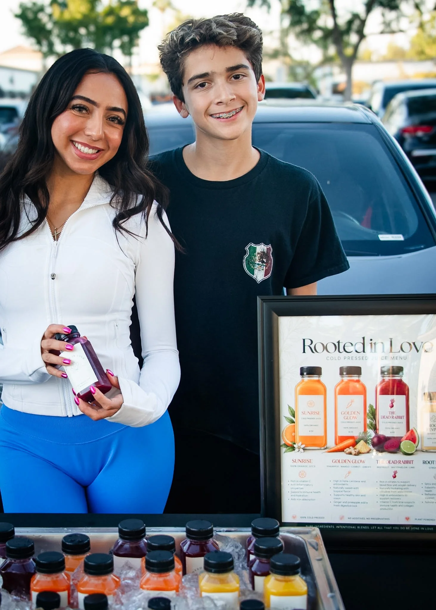 A woman and a teenage boy smiling at a juice stand with bottled juices labeled 'Rooted in Love.' The woman is holding a bottle; the boy is wearing braces, and they are outdoors with cars and trees in the background.