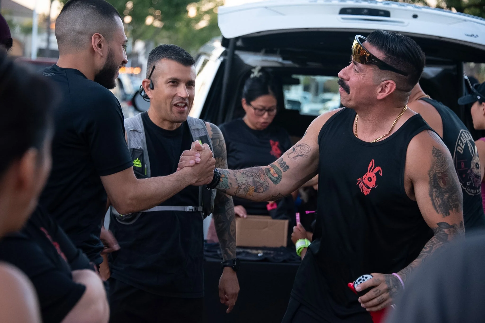 Two men shaking hands and smiling during a gathering outdoors with tents and people in the background, including a woman wearing glasses.