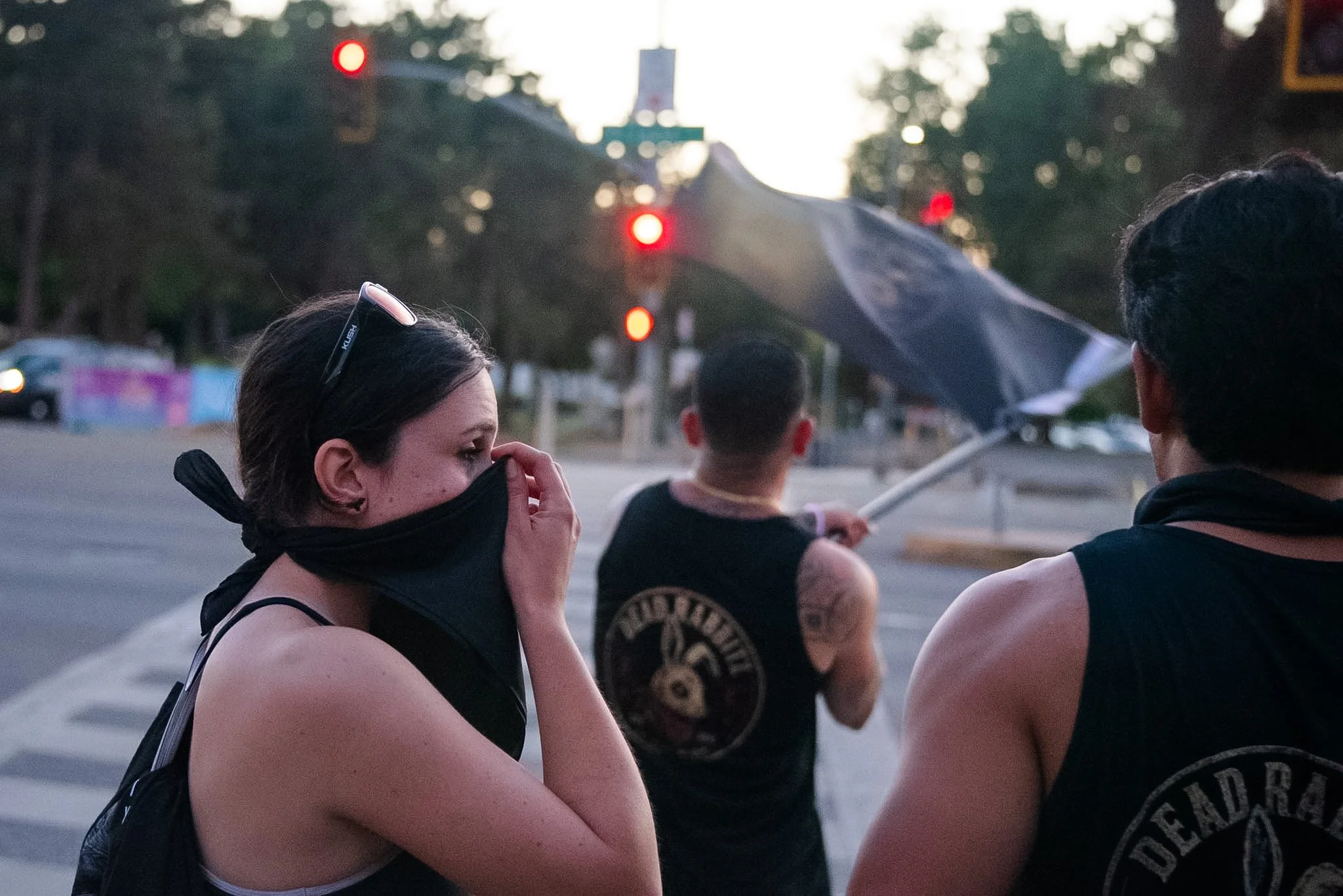 People standing at a street corner during dusk or early evening, with traffic lights and trees in the background. One woman is wearing a face mask, sunglasses on her head, and adjusting her hair. Others are seen from the back, some with tattoos and s