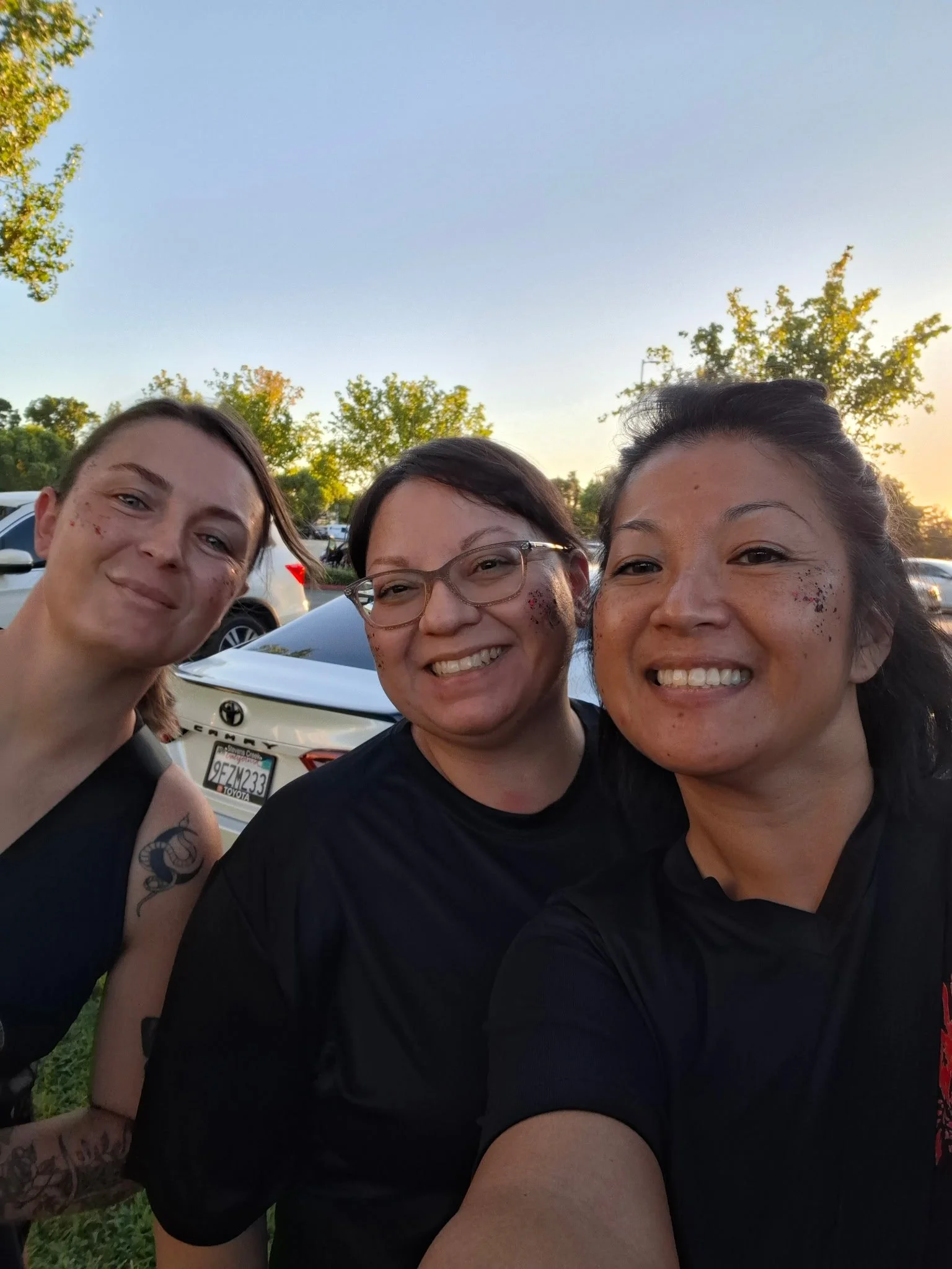 Three women smiling for a selfie outdoors in a parking lot with trees and cars in the background, during sunset.