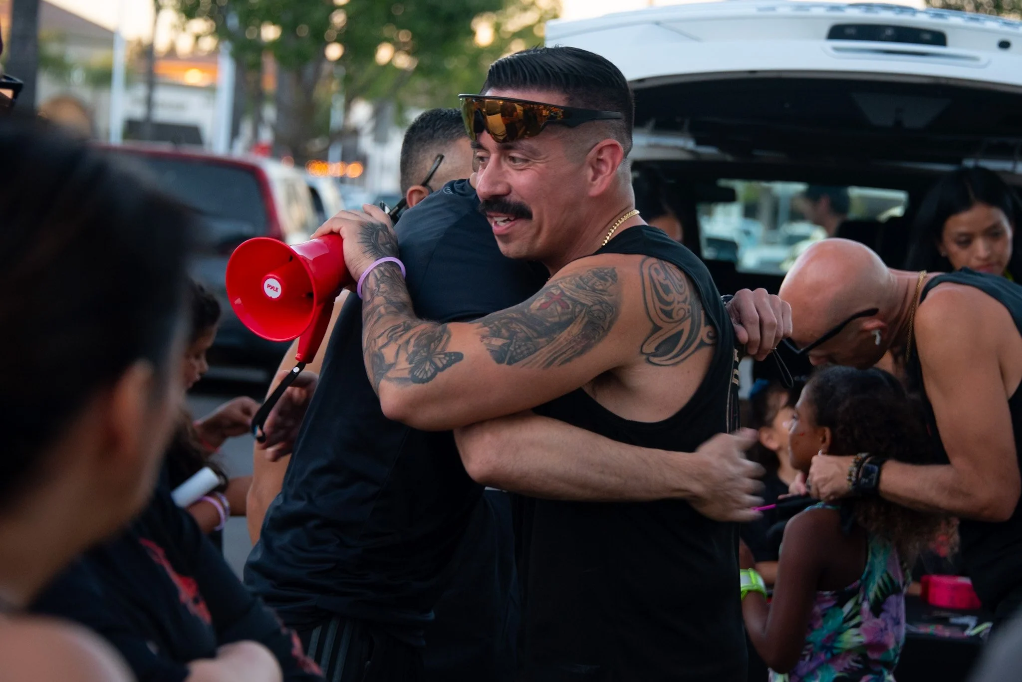 A group of people gather outdoors, with a man with tattoos and sunglasses at the center smiling while hugging someone. Other individuals surround him, some holding objects, and trees are visible in the background.