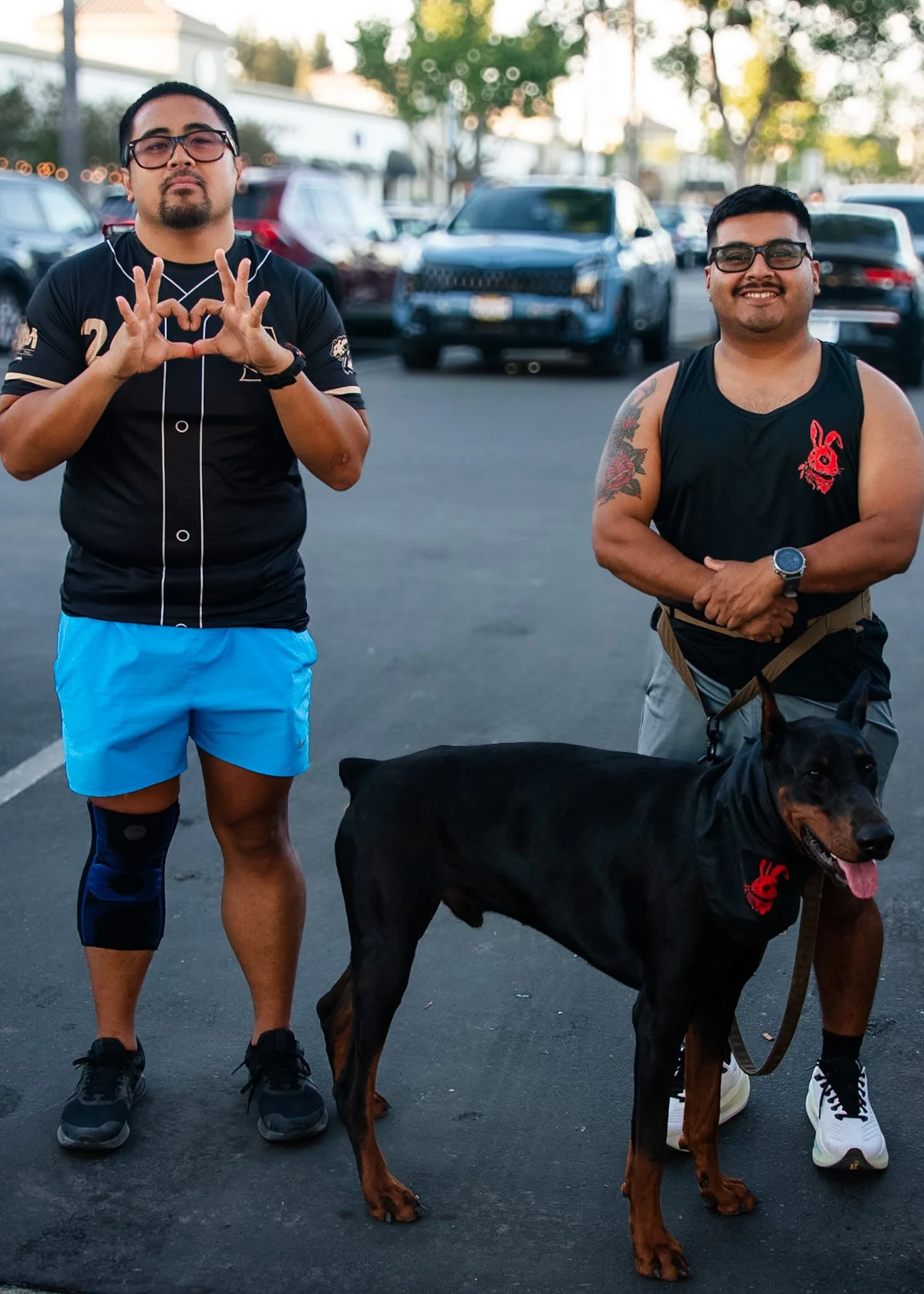 Two men standing outdoors in a parking lot with cars in the background. The man on the left is wearing a black sports jersey with blue shorts, making a heart shape with his hands. The man on the right is smiling, wearing a black sleeveless shirt with