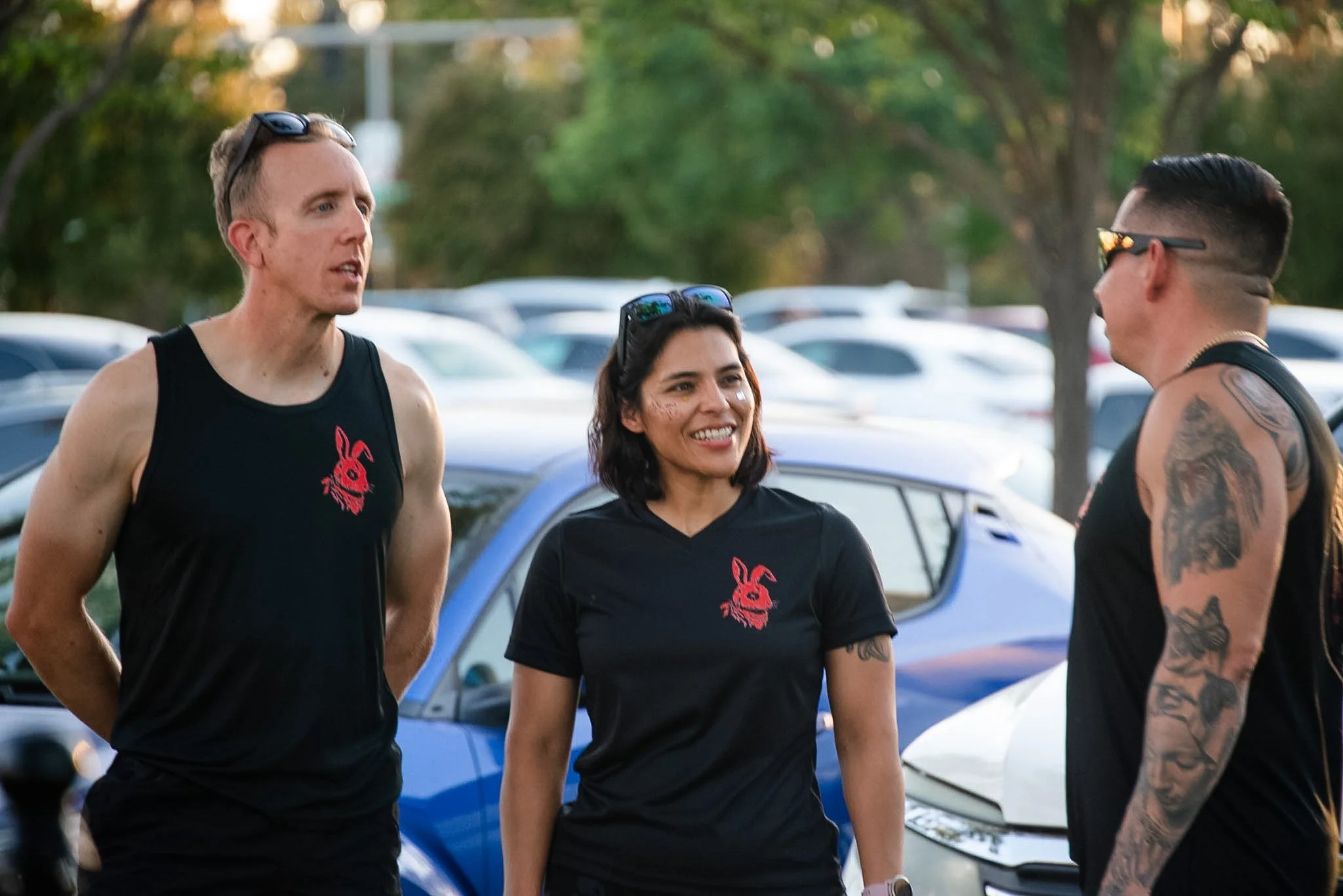 Three people, two men and one woman, standing outdoors and talking. All are wearing black t-shirts with a red bunny logo. The man on the left has sunglasses on his head, and the man on the right has tattoos on his arm and wears sunglasses. The woman 