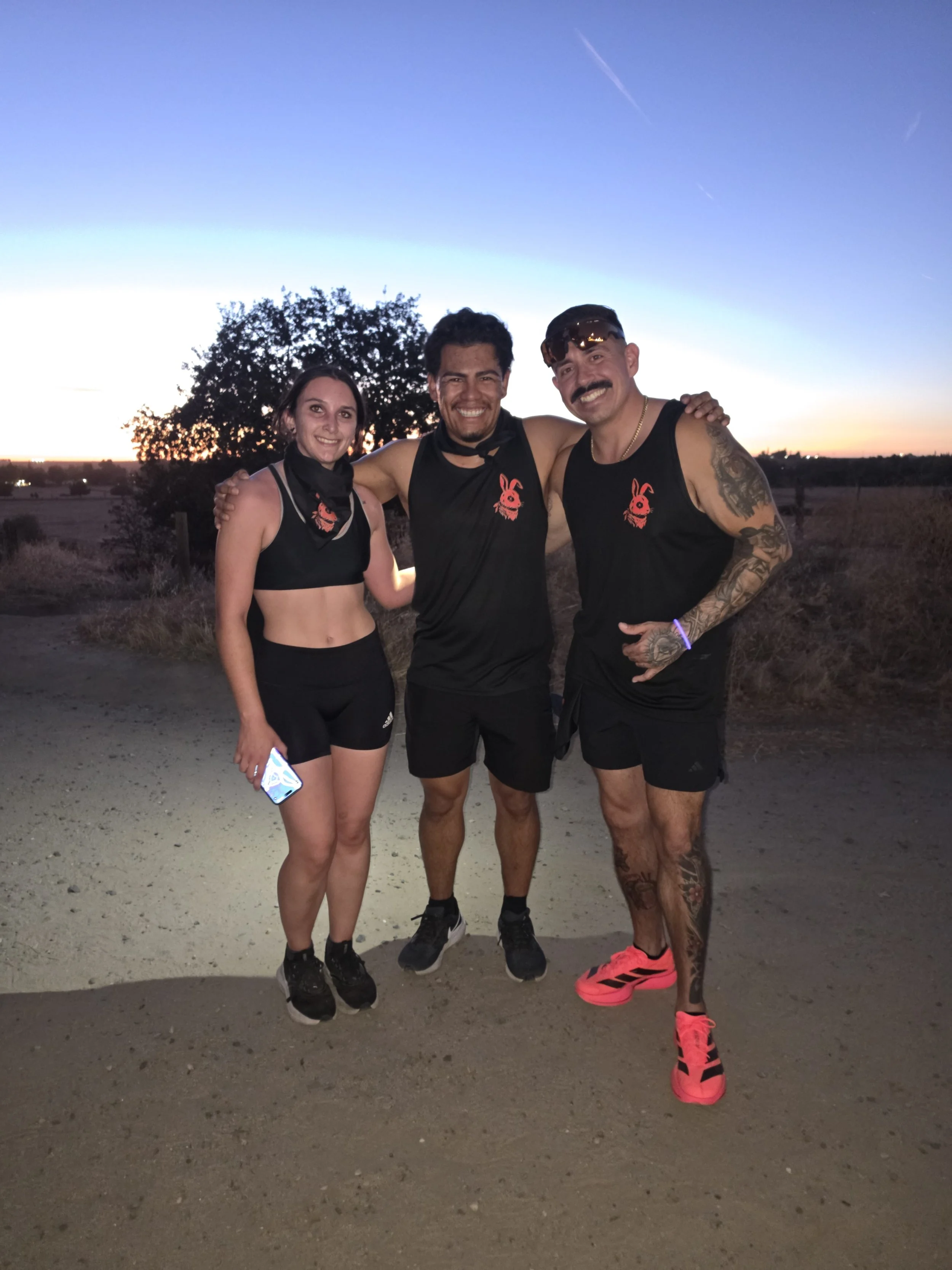 Three people smiling and standing together outdoors during sunset, wearing athletic clothing with matching logos on their shirts, with a tree and horizon in the background.