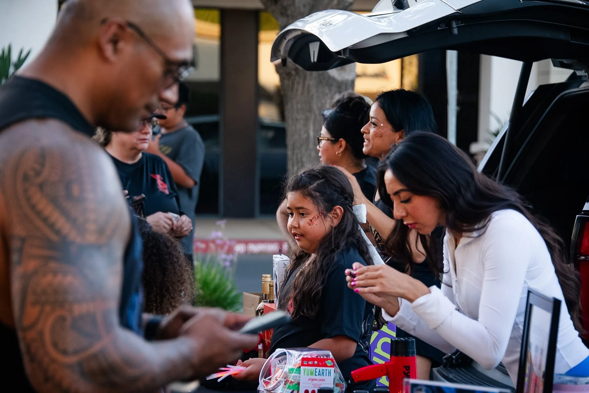 A group of people, including children, gathered around a table at an outdoor event. The table has various items like a box of organic popsicles and a red toy gun. The background shows trees and a parked car.