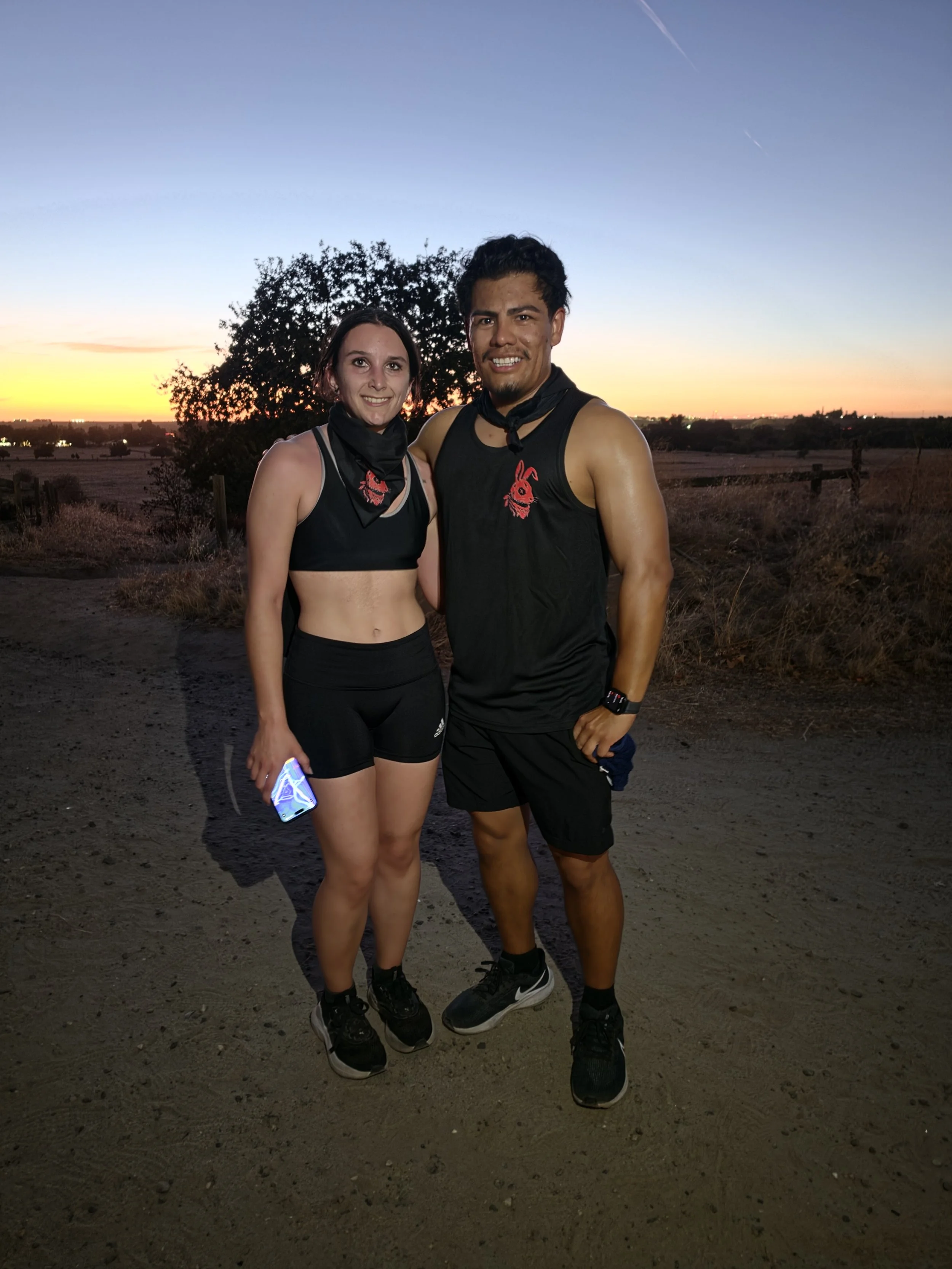 Two athletes in black workout clothes standing outdoors during sunset. They are smiling, with a large tree and sunset sky in the background.