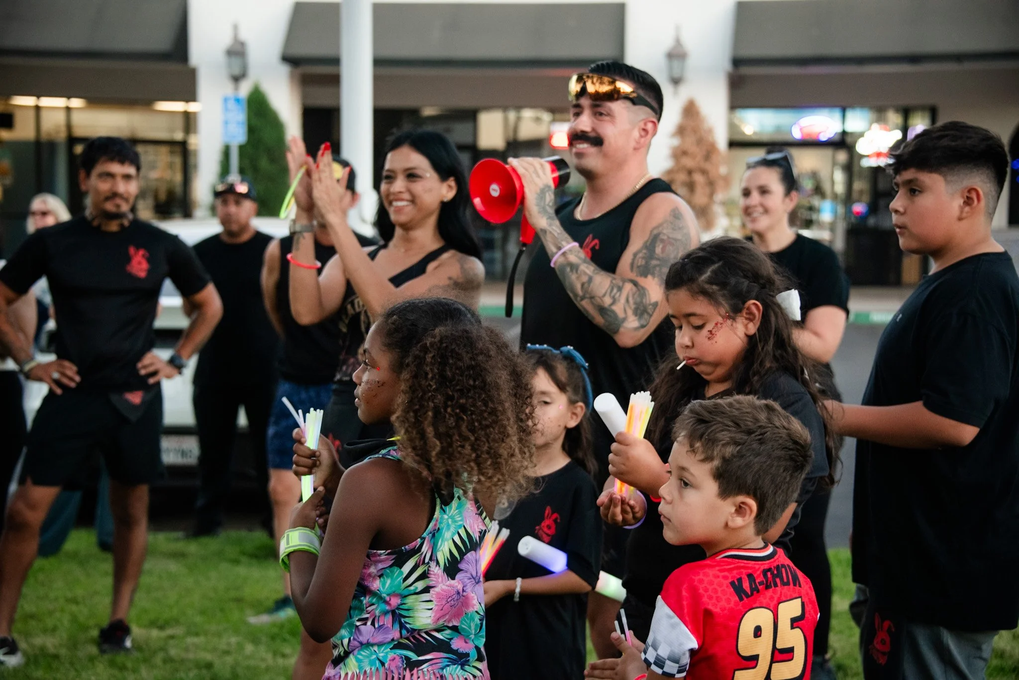 A group of children and adults gathered outdoors at night, holding glow sticks, with a man speaking into a red megaphone. The scene suggests a community event or celebration.